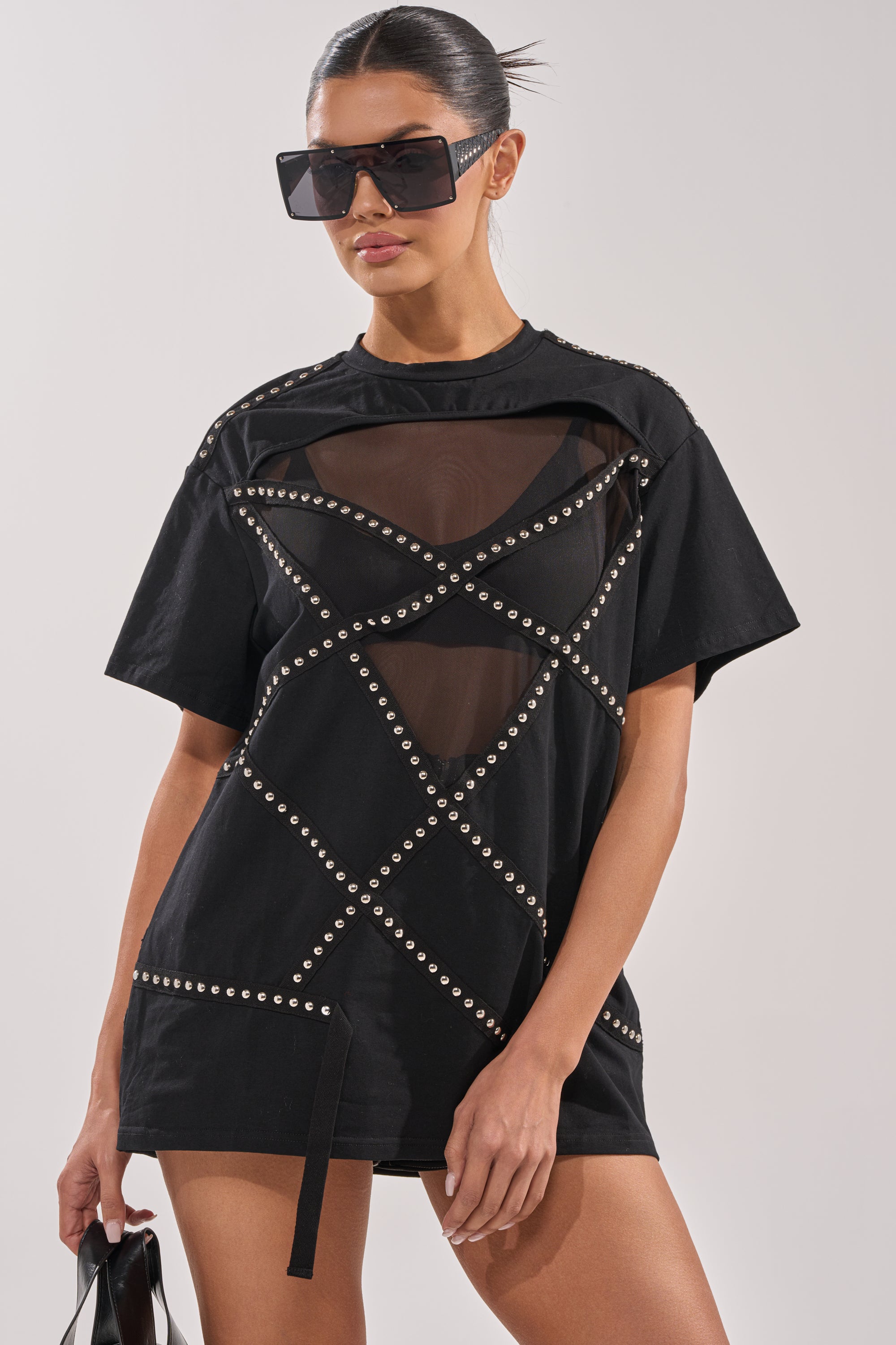 A woman wearing oversized black sunglasses and the MISTA RAJ OVERSIZED TEE with silver studs and mesh paneling stands against a plain background, holding a black bag. 0
