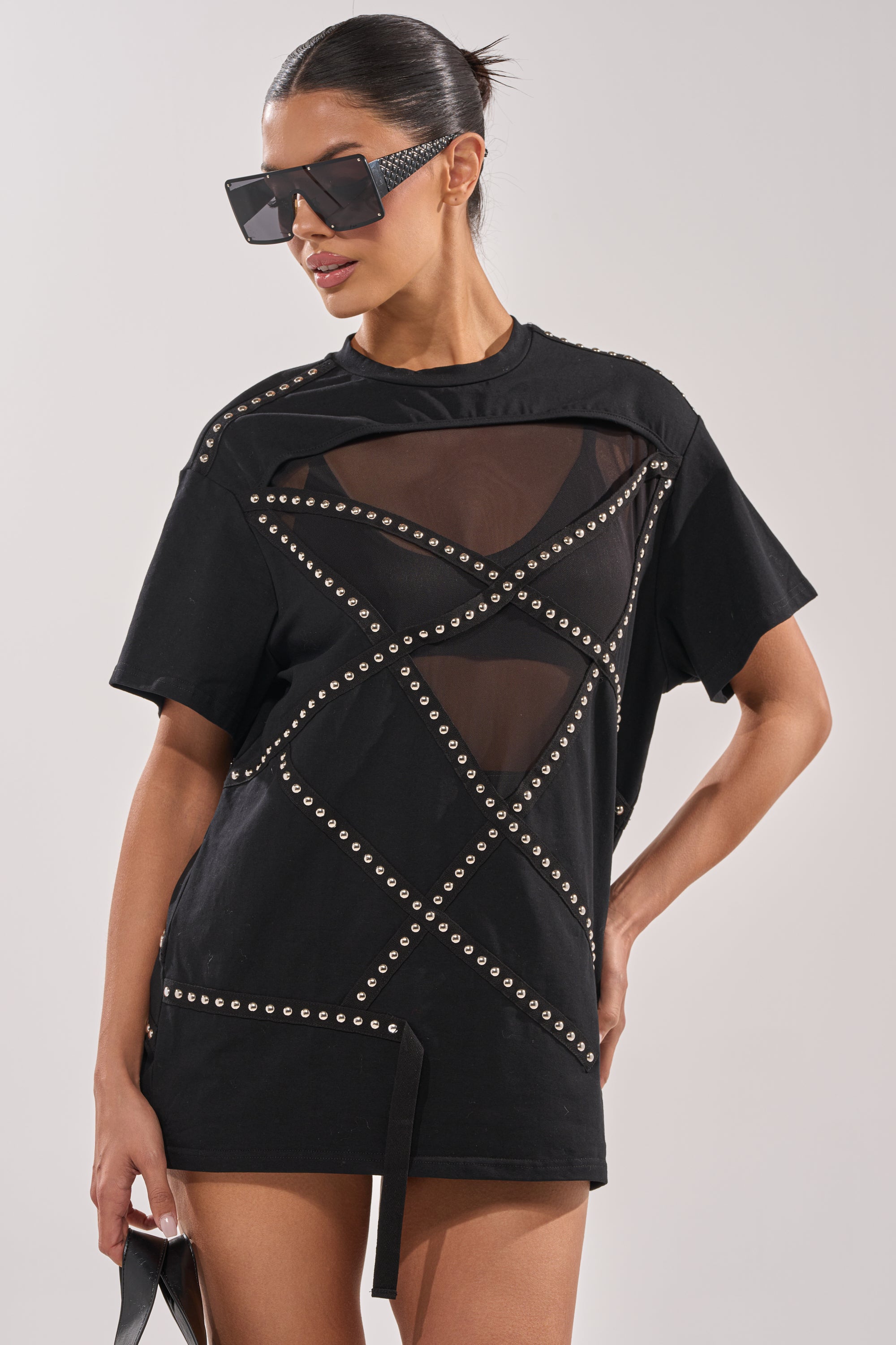 A woman confidently poses against a plain background wearing the MISTA RAJ OVERSIZED TEE, featuring streetwear-inspired black fabric with mesh panels and studded geometric details, plus oversized black sunglasses and her hair pulled back. 1