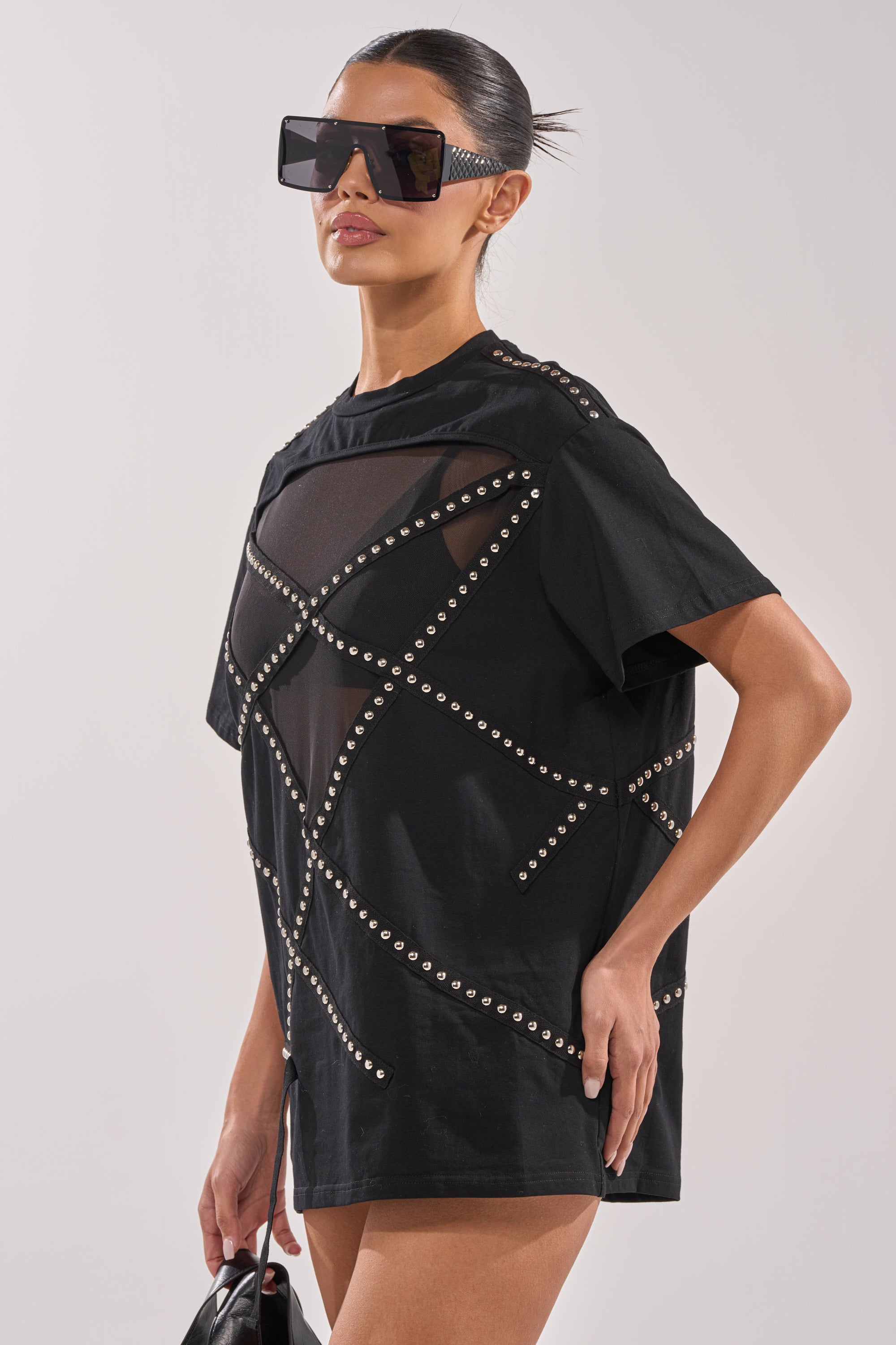 A woman in MISTA RAJ OVERSIZED TEE with mesh panels and stud embellishments, oversized square sunglasses, and a small black bag poses against a plain background. 2