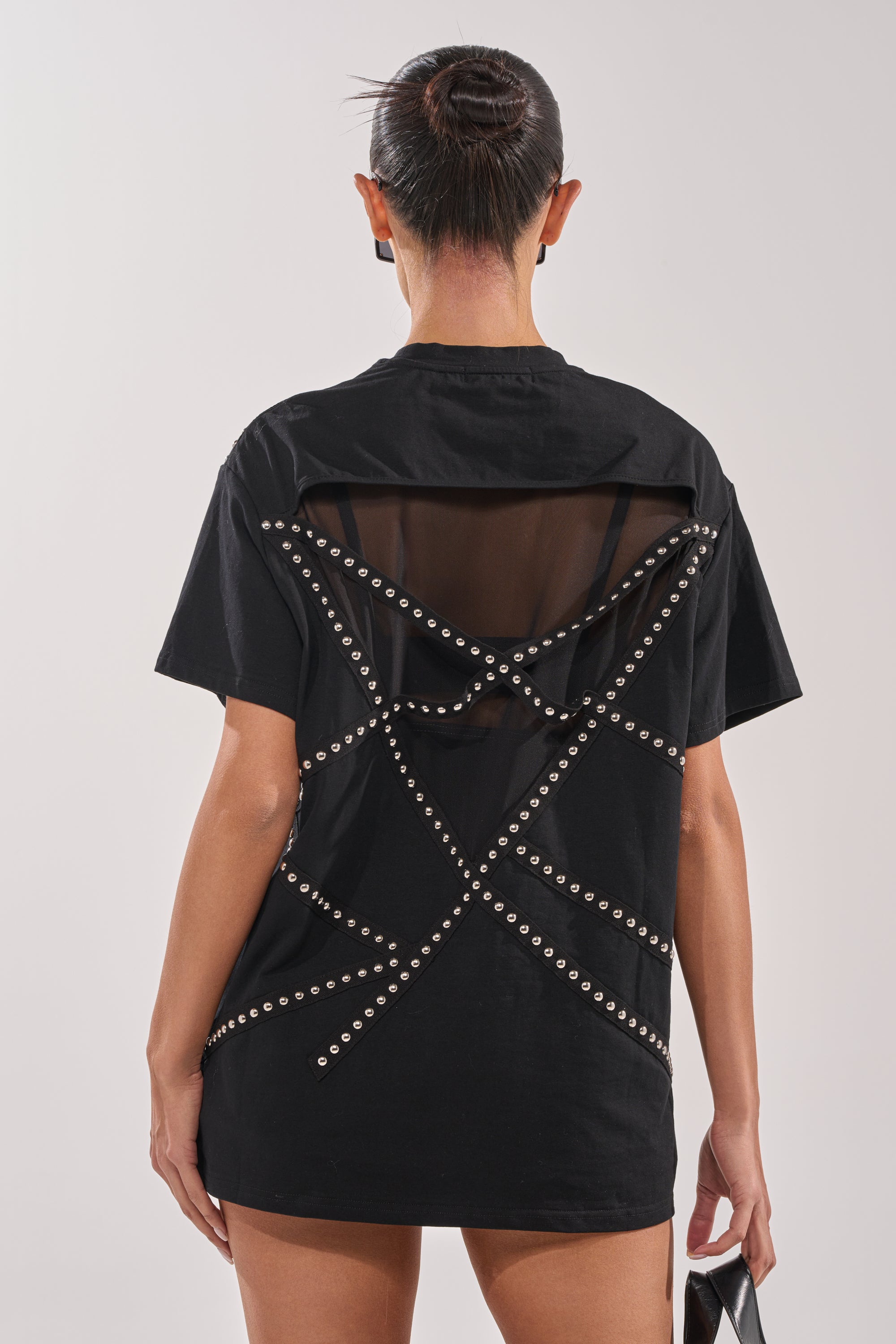 A person with a neat bun wears the MISTA RAJ OVERSIZED TEE in black, featuring sheer mesh back paneling and crisscross silver studs. The front isn’t visible, and the background is plain and light-colored. 3