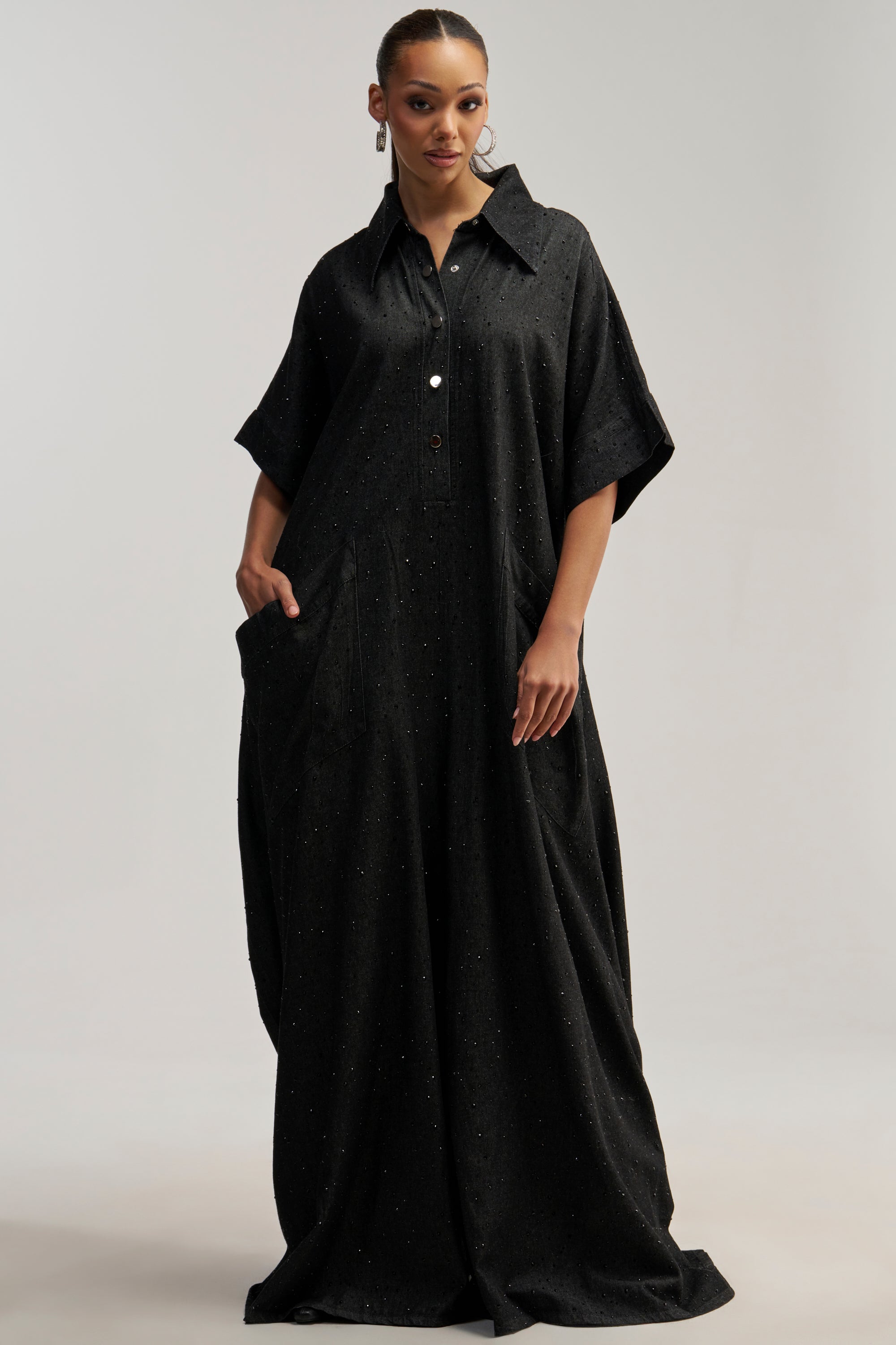 A woman stands with quiet confidence against a plain background, wearing the GINA DENIM RHINESTONE JUMPSUIT IN BLACK—a loose-fitting, floor-length black jumpsuit featuring short sleeves, a crisp collar, front buttons, and pockets—offering a bold new take on statement streetwear.