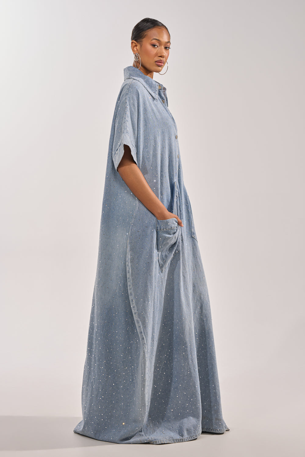 A woman poses in profile wearing the GINA DENIM RHINESTONE JUMPSUIT—a long, light blue, short-sleeve piece with sparkling embellishments, a loose fit, and pockets. Her hair is pulled back as she stands against a plain background. 8
