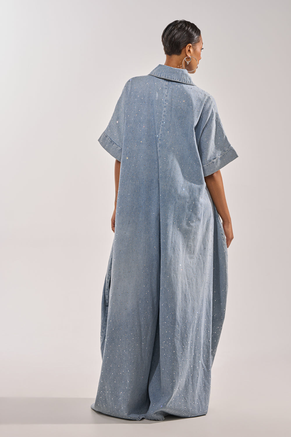 A person with short hair is seen from behind wearing the GINA DENIM RHINESTONE JUMPSUIT, a loose, light blue wide-leg jumpsuit with short sleeves, subtle white speckles, a wide collar, and a graceful floor-length silhouette. 10