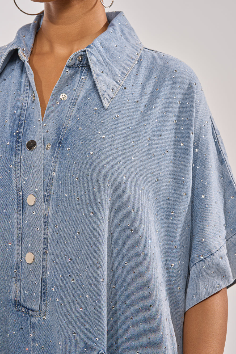 Close-up of a person wearing the GINA DENIM RHINESTONE JUMPSUIT, featuring light blue denim, rhinestone accents, metal snap buttons, a pointed collar, and rolled short sleeves. Styled with large hoop earrings for a trendy look. 11