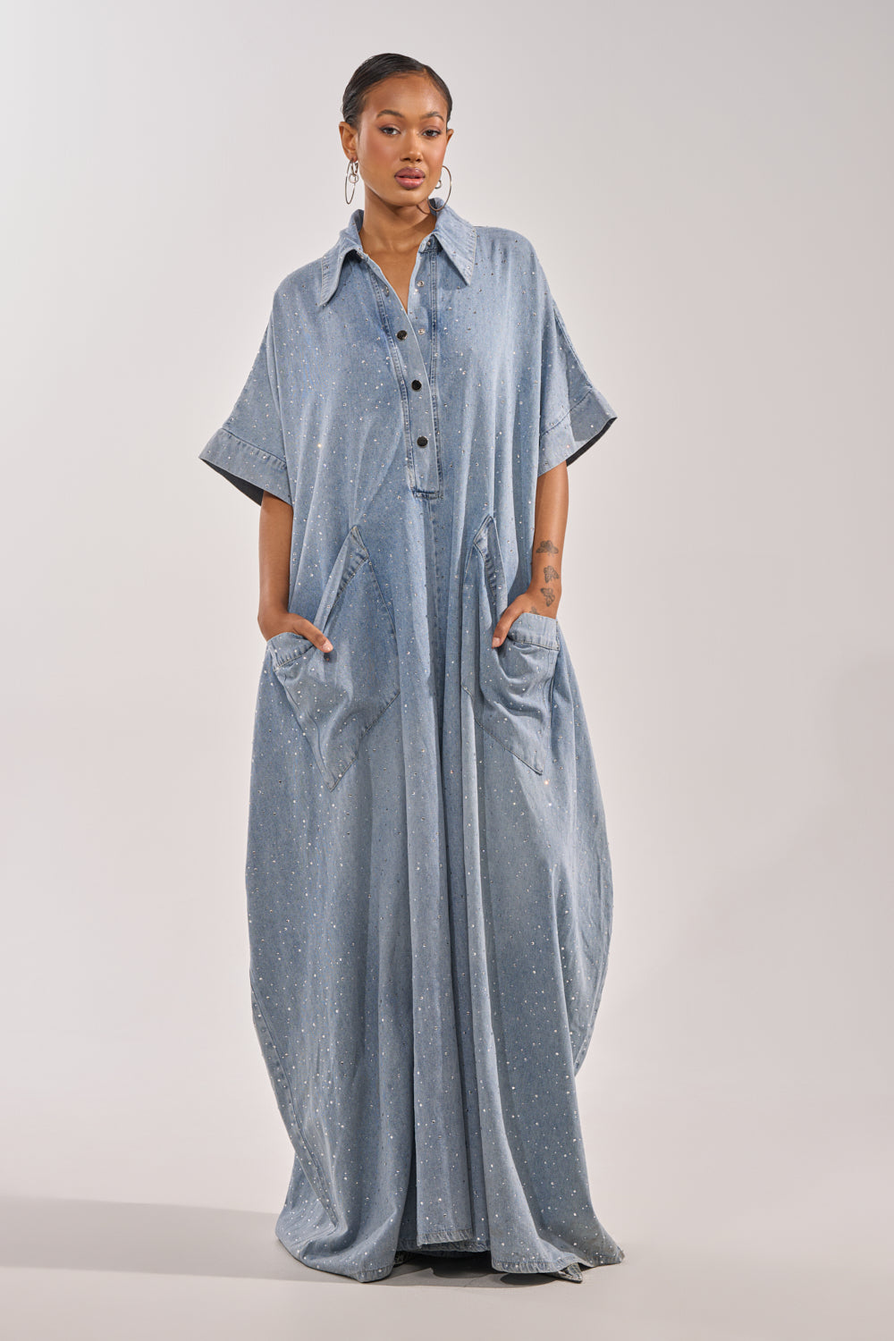 A woman wears the GINA DENIM RHINESTONE JUMPSUIT, a loose-fitting, light blue denim outfit with short sleeves, large front pockets, and subtle sparkling rhinestone accents, standing with her hands in her pockets against a plain background. 1