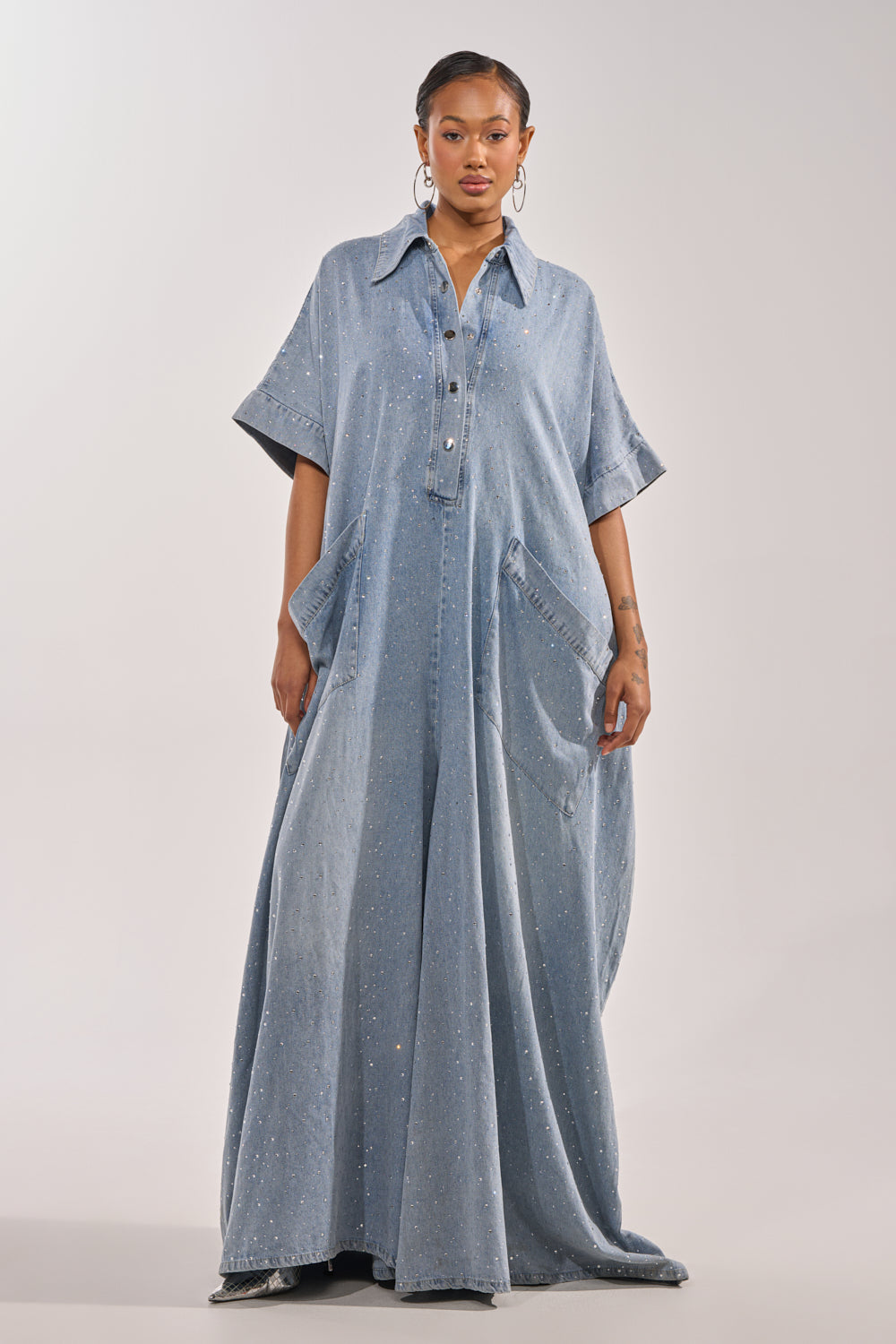 A woman faces forward in the GINA DENIM RHINESTONE JUMPSUIT, a loose, light blue denim piece with short sleeves and silver buttons. She wears hoop earrings, her hair pulled back, against a plain light gray background. 2