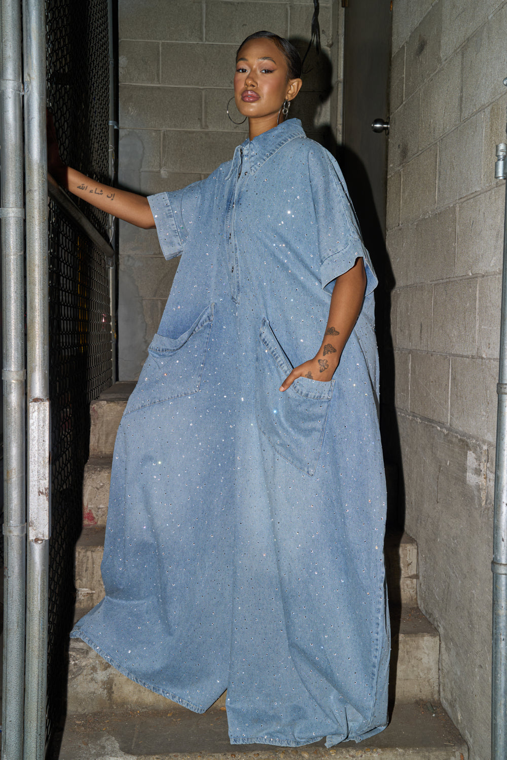 A woman with short hair, tattoos, and earrings stands on concrete steps in an industrial setting, wearing the GINA DENIM RHINESTONE JUMPSUIT—a bold, oversized piece featuring rhinestones, short sleeves, and large pockets. 4