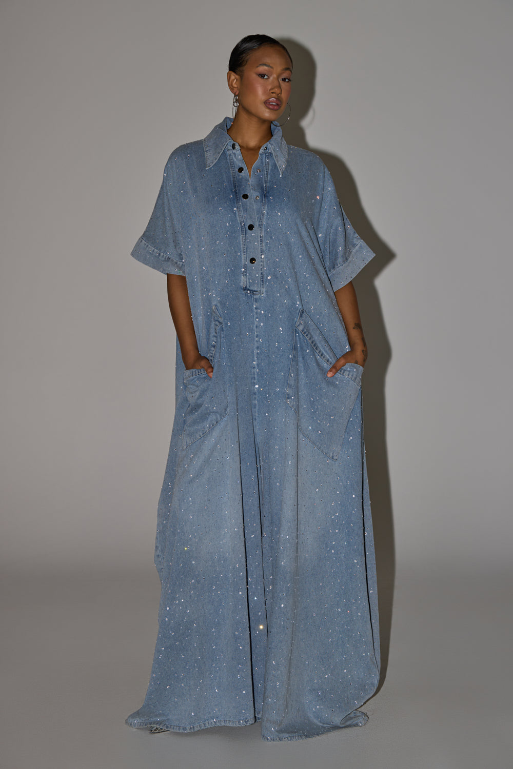 A woman with short hair poses against a plain background wearing the GINA DENIM RHINESTONE JUMPSUIT, a loose, floor-length denim piece with short sleeves, large pockets, and sparkling rhinestone embellishments. Her hands rest in her pockets. 6