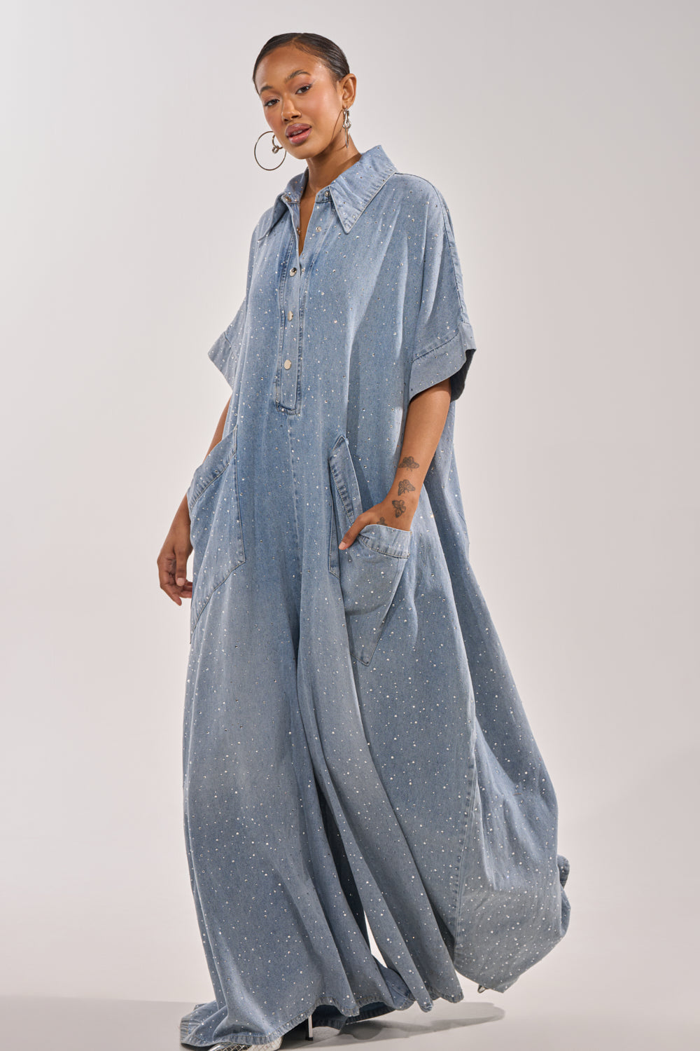 A woman wears the GINA DENIM RHINESTONE JUMPSUIT, a relaxed, light blue denim piece with short sleeves, large pockets, and a collar. The wide-leg design and subtle speckled pattern make it stand out against a plain background. 7