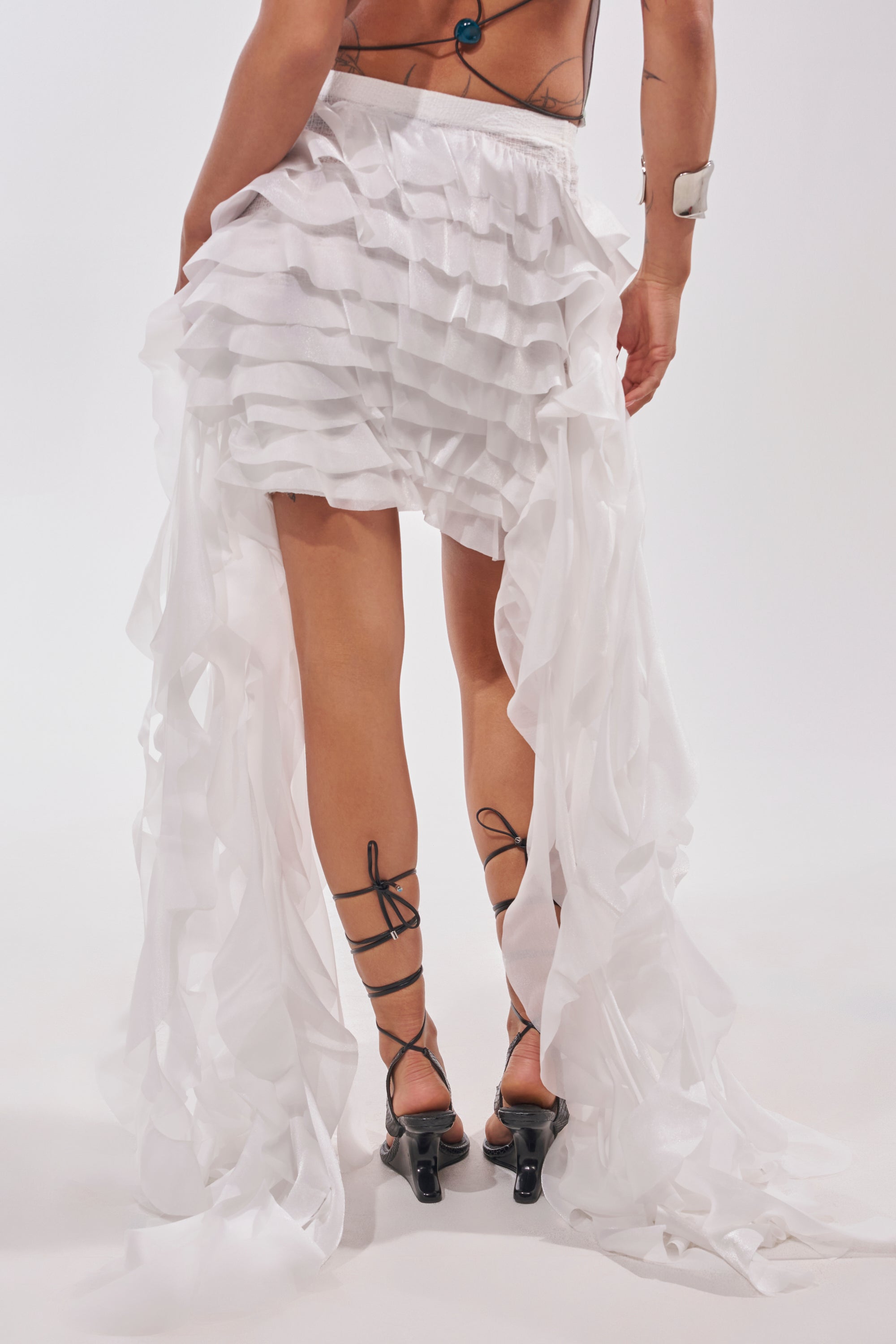 Model wears the TAKE ME AWAY MINI SKIRT, a ruffled white mini with trailing fabric strips, paired with black strappy heels and a blue halter top. Plain white background; image captures a back view from the waist down. 3