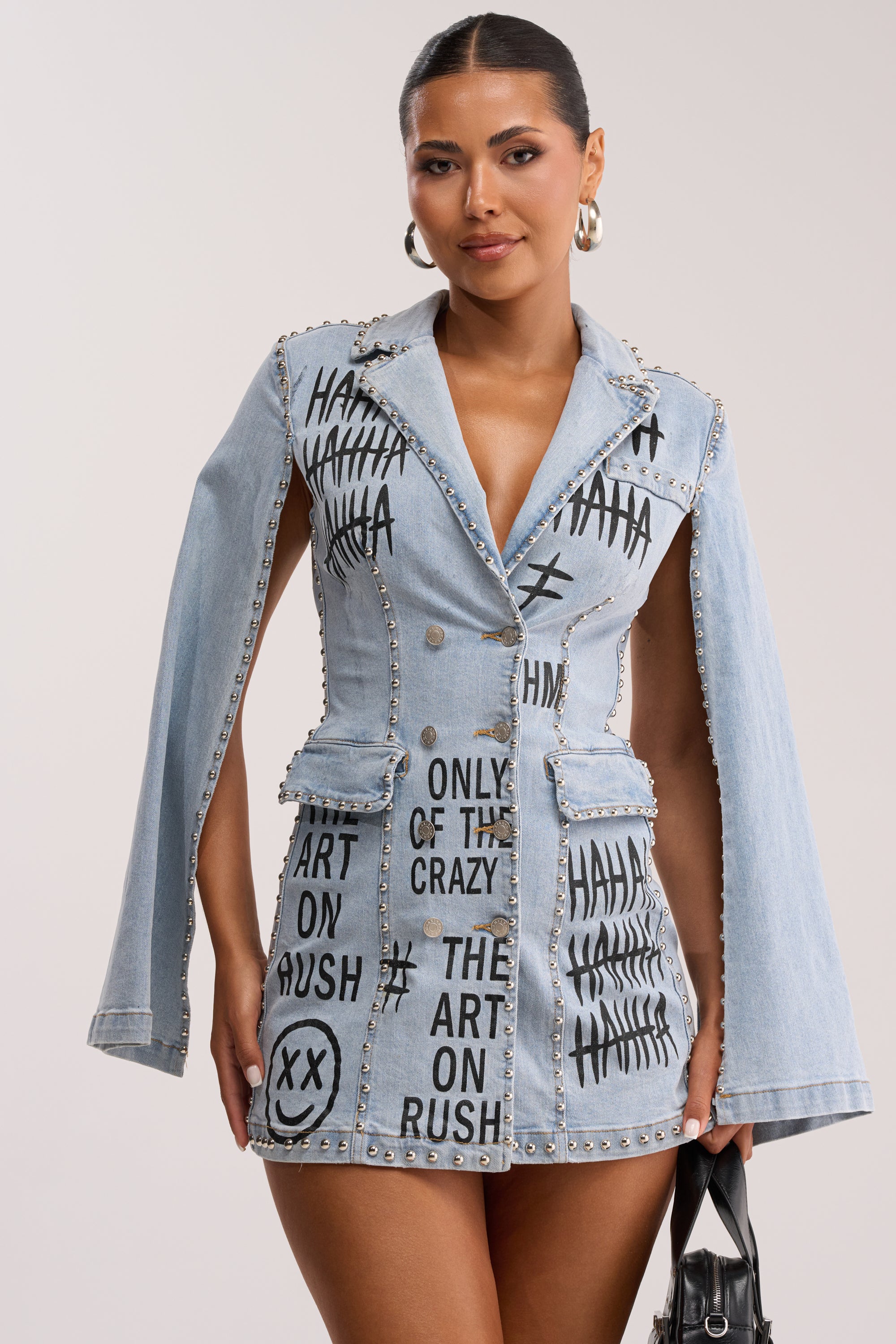 A woman wears the MARK MY WORDS GRAPHIC STUDDED CAPE DRESS, a blazer-style embellished denim mini dress with silver studs and black graffiti text. She pairs it with a black handbag, slicked-back hair, and hoop earrings.