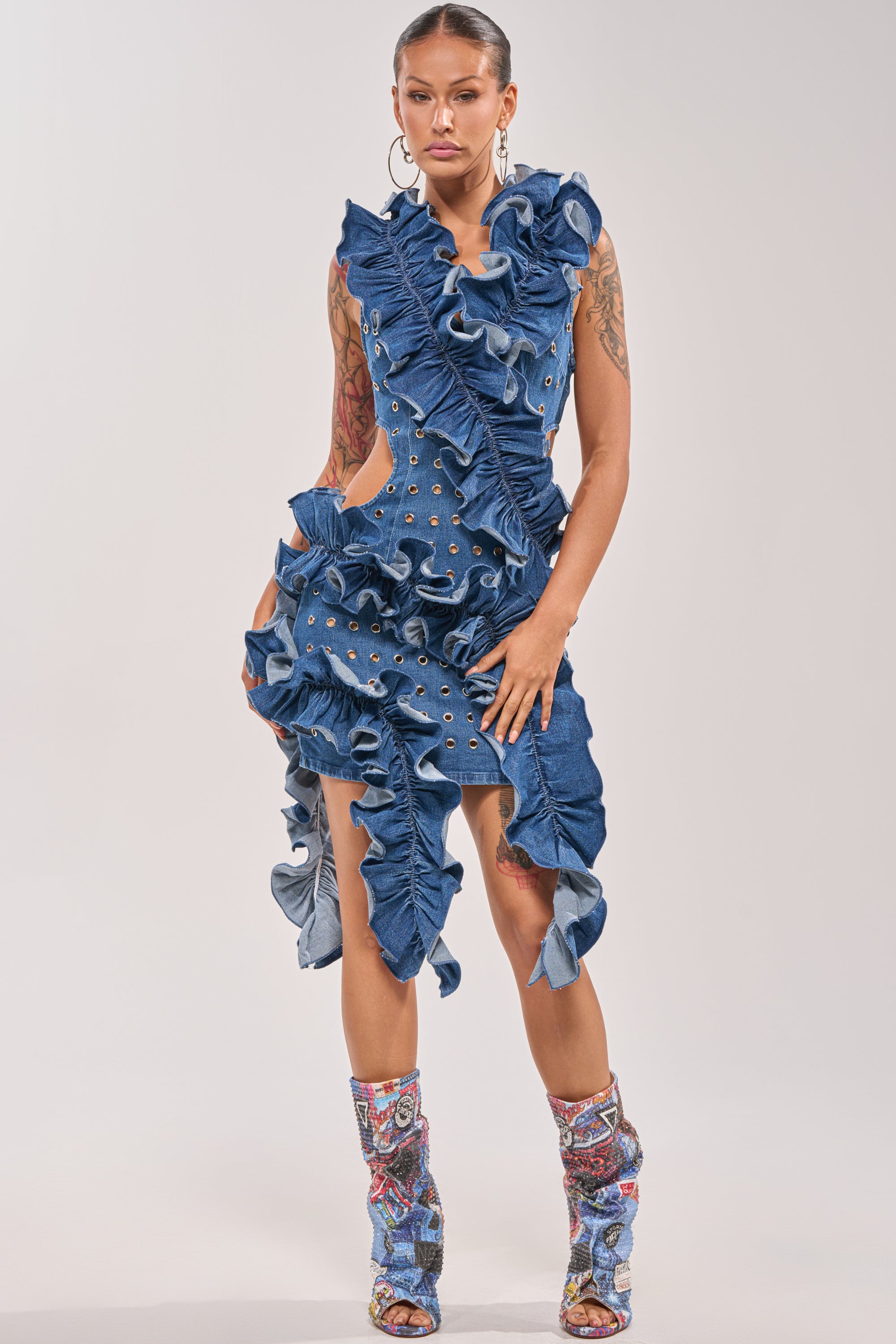 A woman stands confidently in the LINGERING EYES FLOCKED DENIM MINI DRESS, featuring ruffles and grommets, paired with colorful knee-high boots. She showcases arm tattoos and large hoops against a plain light gray background. 0