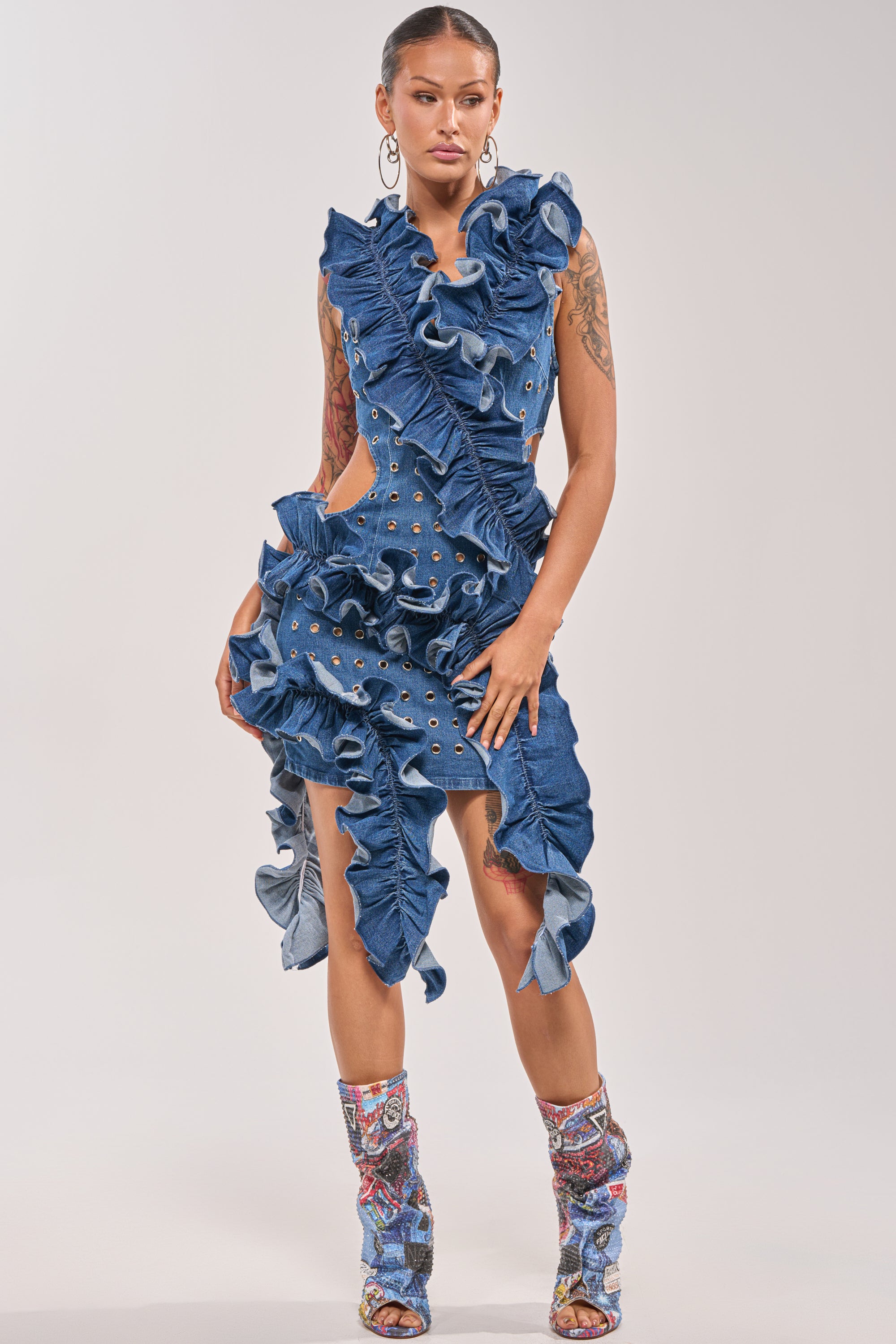 A woman exudes confidence in the LINGERING EYES FLOCKED DENIM MINI DRESS, featuring cutouts, ruffles, and grommets. Paired with matching boots and hoop earrings, her tattoos and sleek hair complete a bold festival-ready look against a plain backdrop. 1