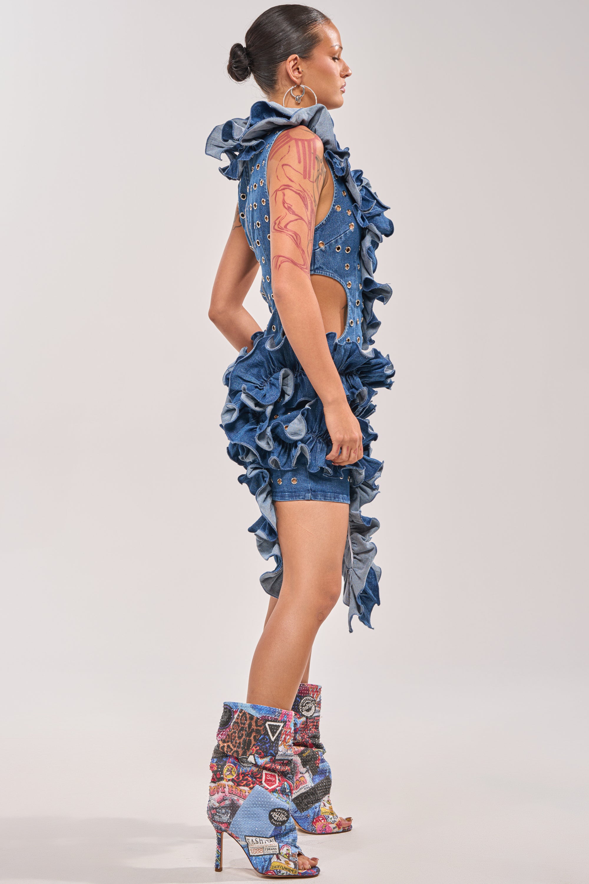 A woman in profile wears the LINGERING EYES FLOCKED DENIM MINI DRESS with side cutouts, grommets, and high-heeled patchwork boots. Her slicked-back bun and bold pink abstract tattoo complete her edgy streetwear look. 2