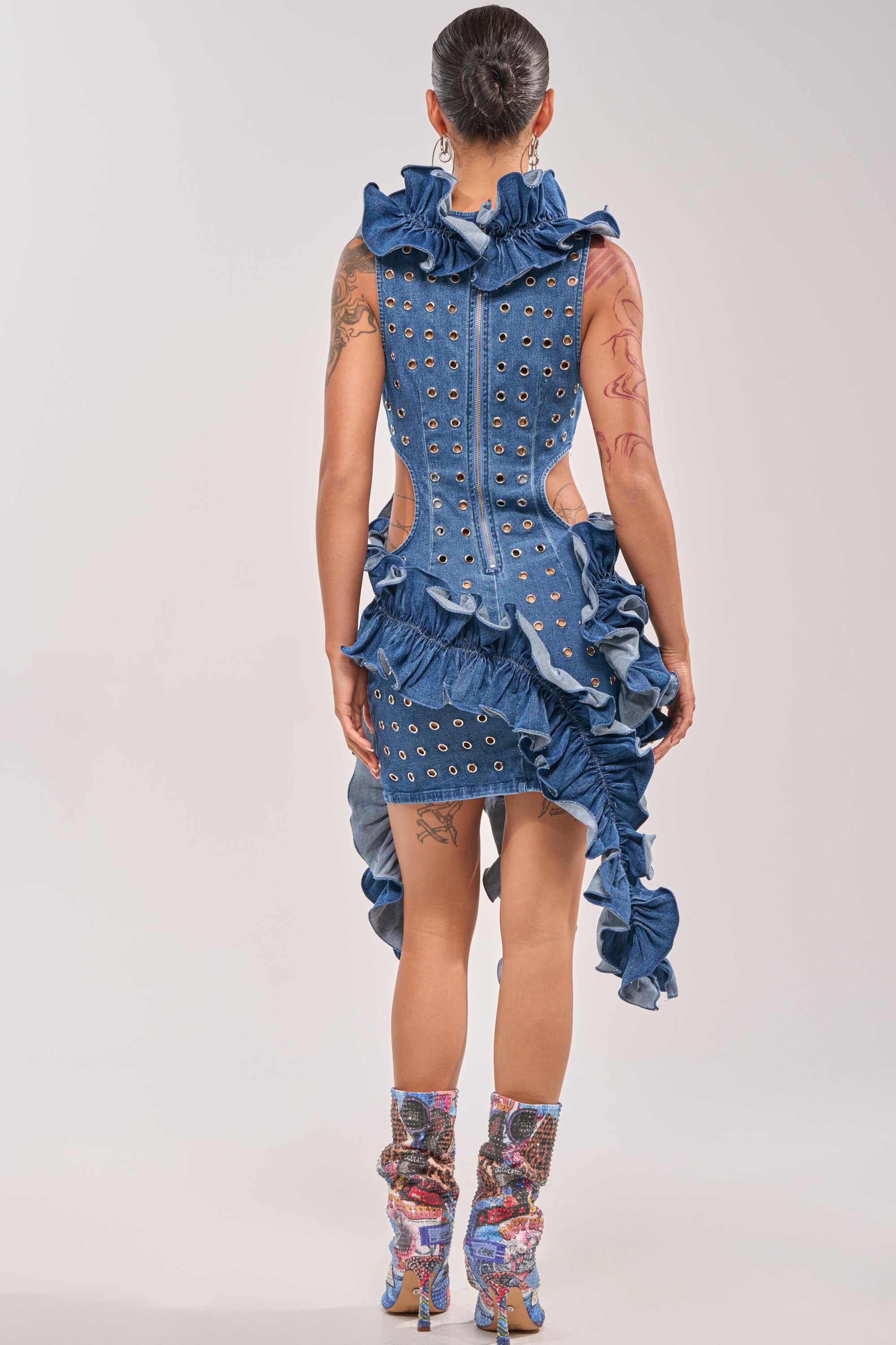 A person faces away in the LINGERING EYES FLOCKED DENIM MINI DRESS, featuring metal eyelets, ruffles, an asymmetrical hem, and cutouts—styled with patterned heeled boots. Visible arm and leg tattoos add a bold streetwear edge. 4