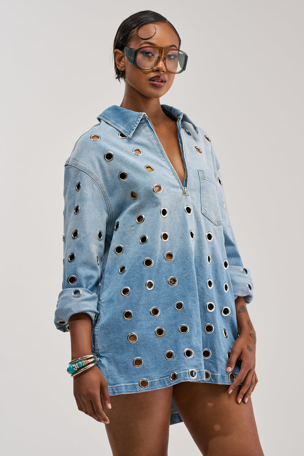 Sporting the COOL GIRL OVERSIZED GROMMET SHIRT DRESS IN DENIM and large sunglasses, a person stands confidently against a plain light background.