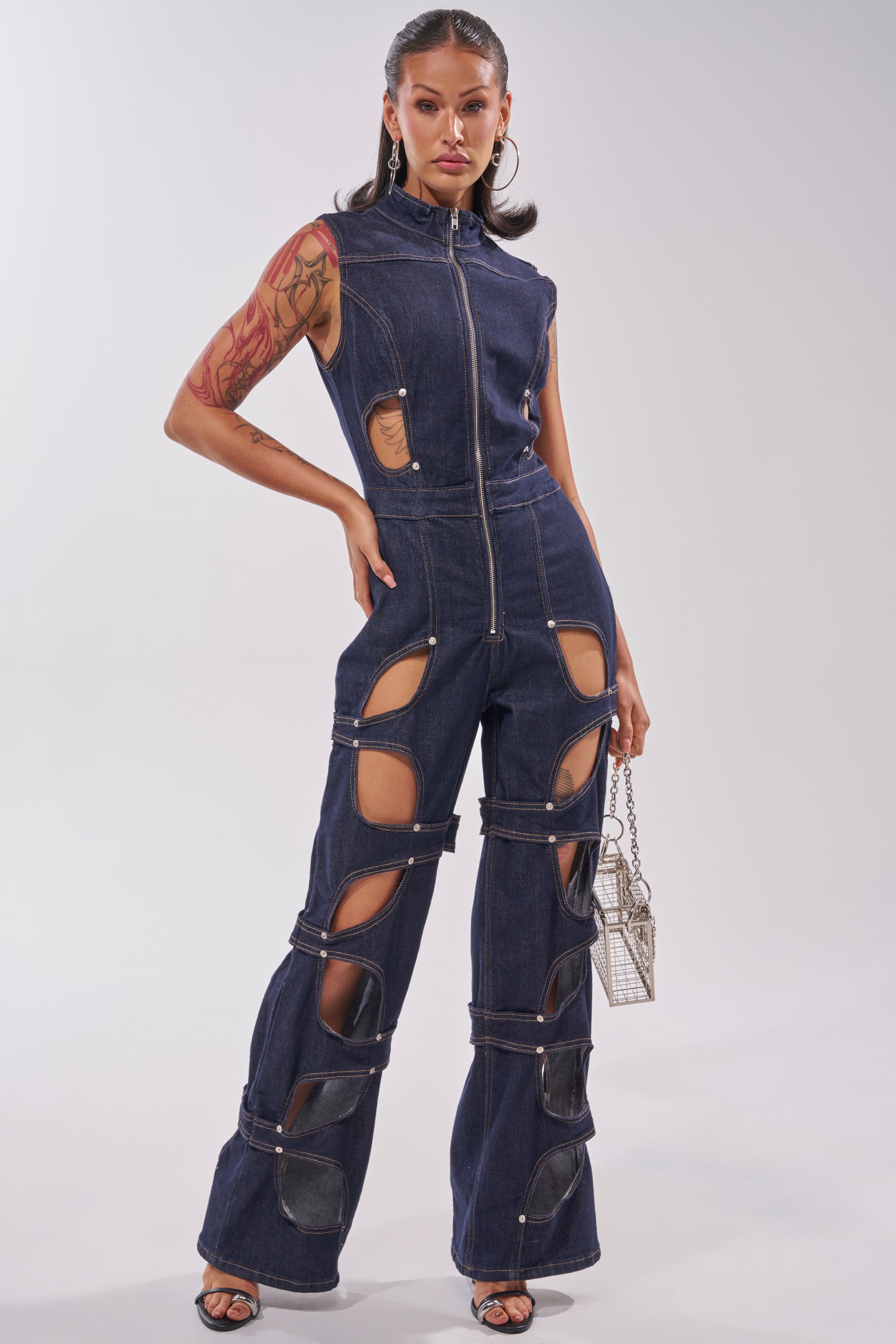 A woman poses confidently against a plain light background wearing the BRING THE ENERGY CUT OUT DENIM JUMPSUIT, paired with black heeled sandals and a metallic handbag. 0