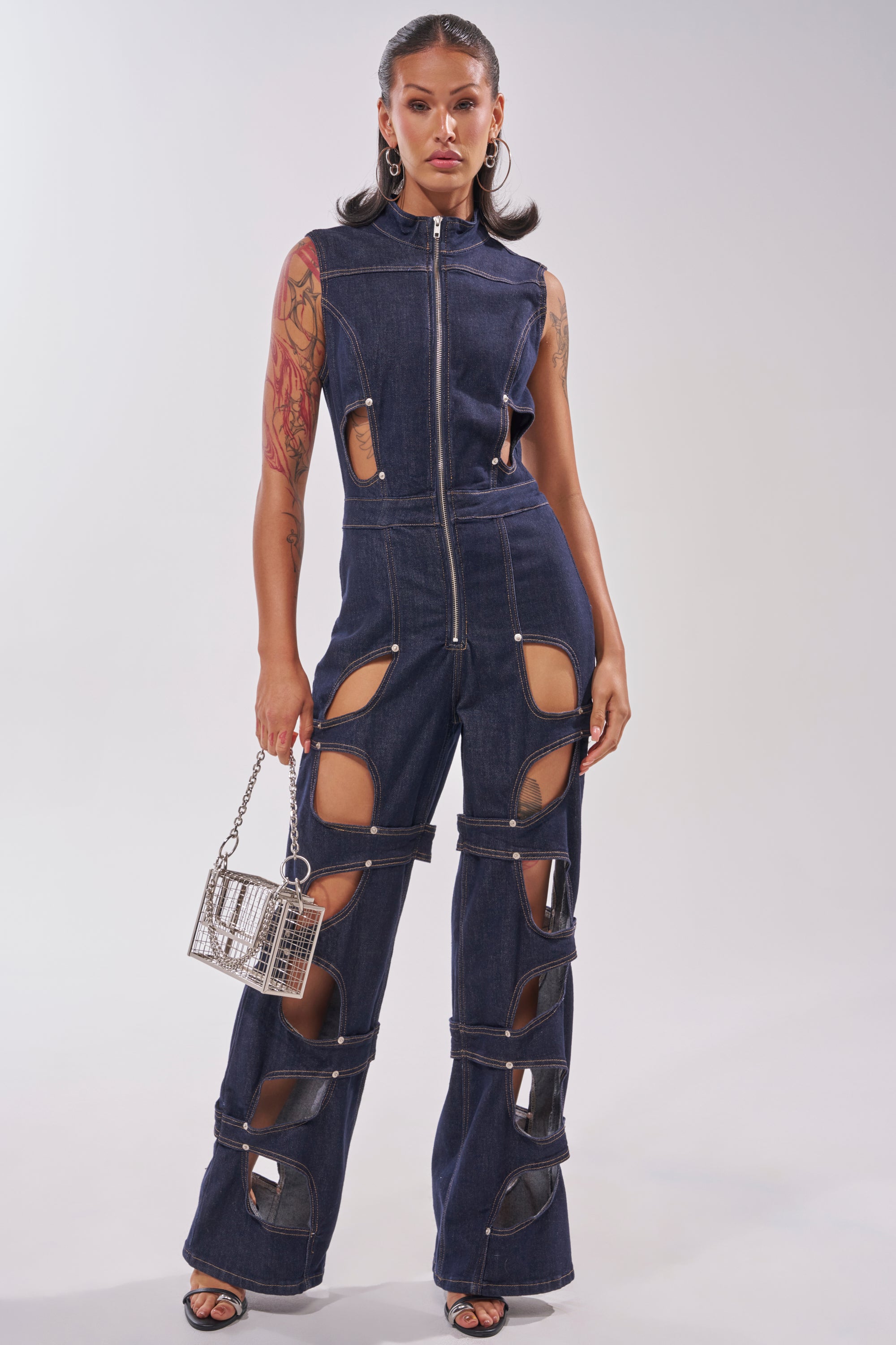 A woman poses confidently in the BRING THE ENERGY CUT OUT DENIM JUMPSUIT, featuring dark-wash denim and flared legs. She accessorizes with hoop earrings, high heels, a silver handbag, and shows off her arm tattoos. 1