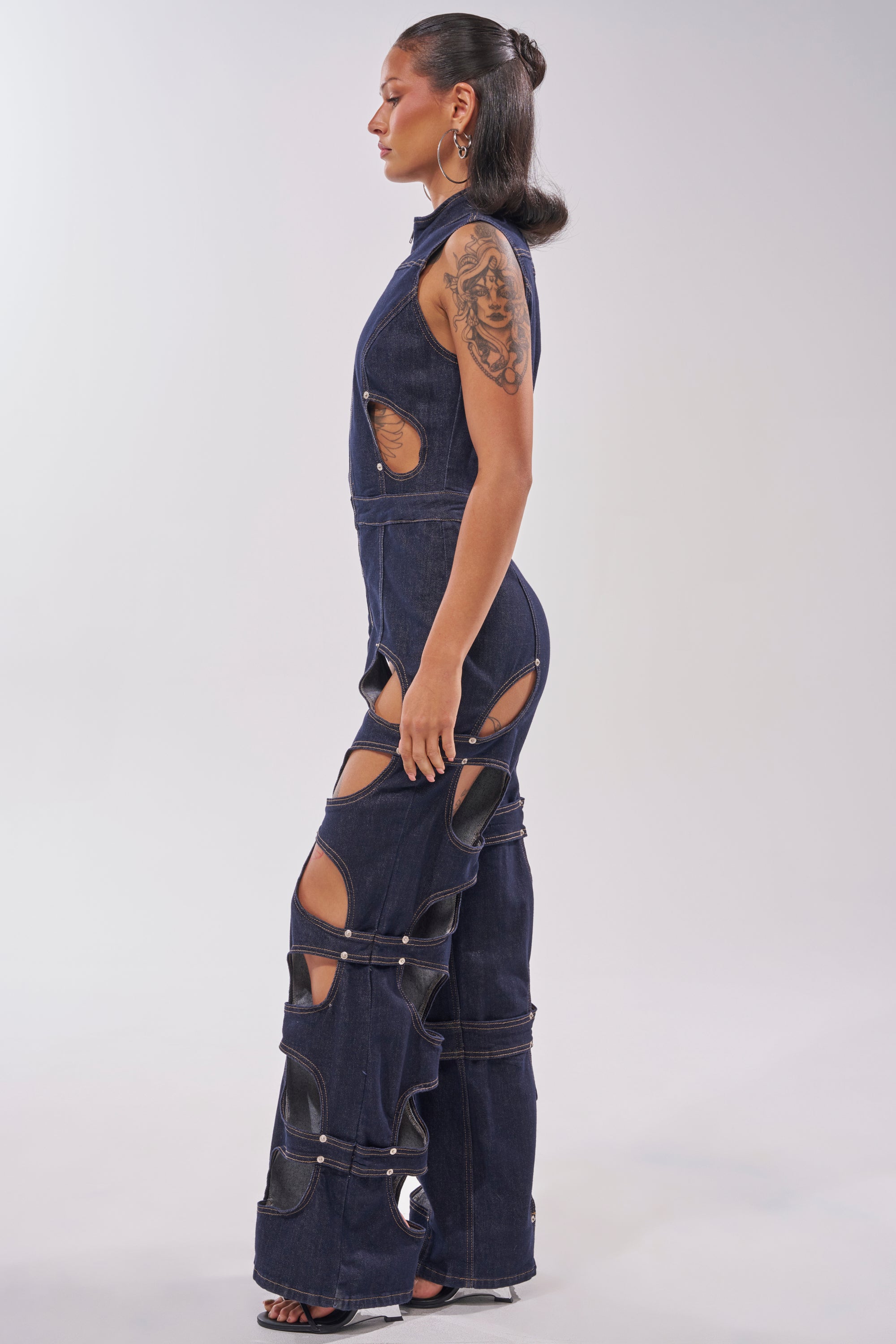 A woman in profile wears the BRING THE ENERGY CUT OUT DENIM JUMPSUIT, featuring dark-wash denim, multiple circular cutouts on the torso, and flared legs. She has a tattoo on her upper arm and her hair is styled in a sleek low ponytail. 2