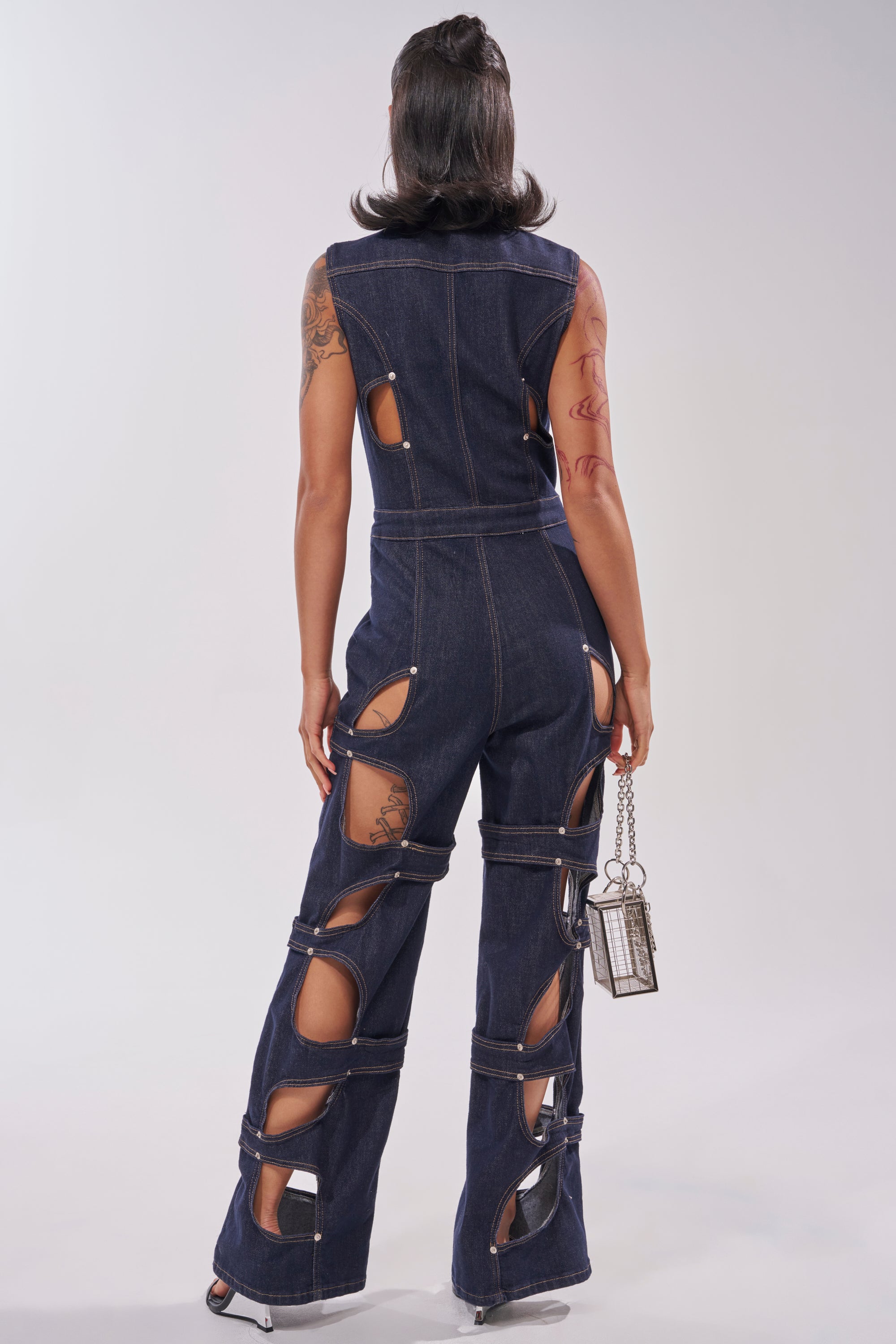Wearing the BRING THE ENERGY CUT OUT DENIM JUMPSUIT, a person stands facing away, showing off its sleeveless design, flared legs, and bold back, leg, and side cutouts. They carry a small silver bag and display arm tattoos. 3