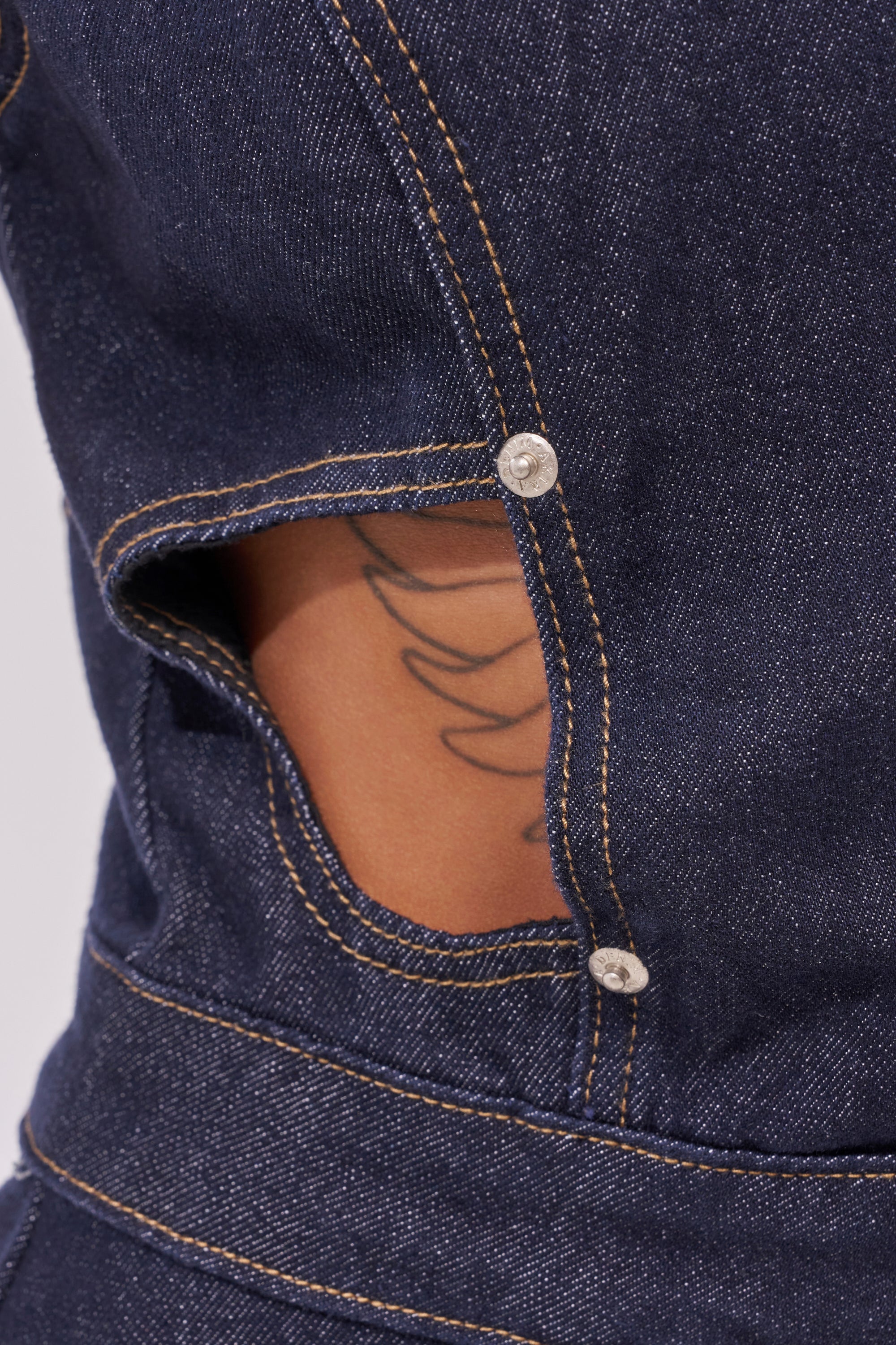 Close-up of the BRING THE ENERGY CUT OUT DENIM JUMPSUIT in dark wash with yellow stitching, featuring a cutout secured by silver buttons that reveals skin and part of a black abstract tattoo underneath. 4