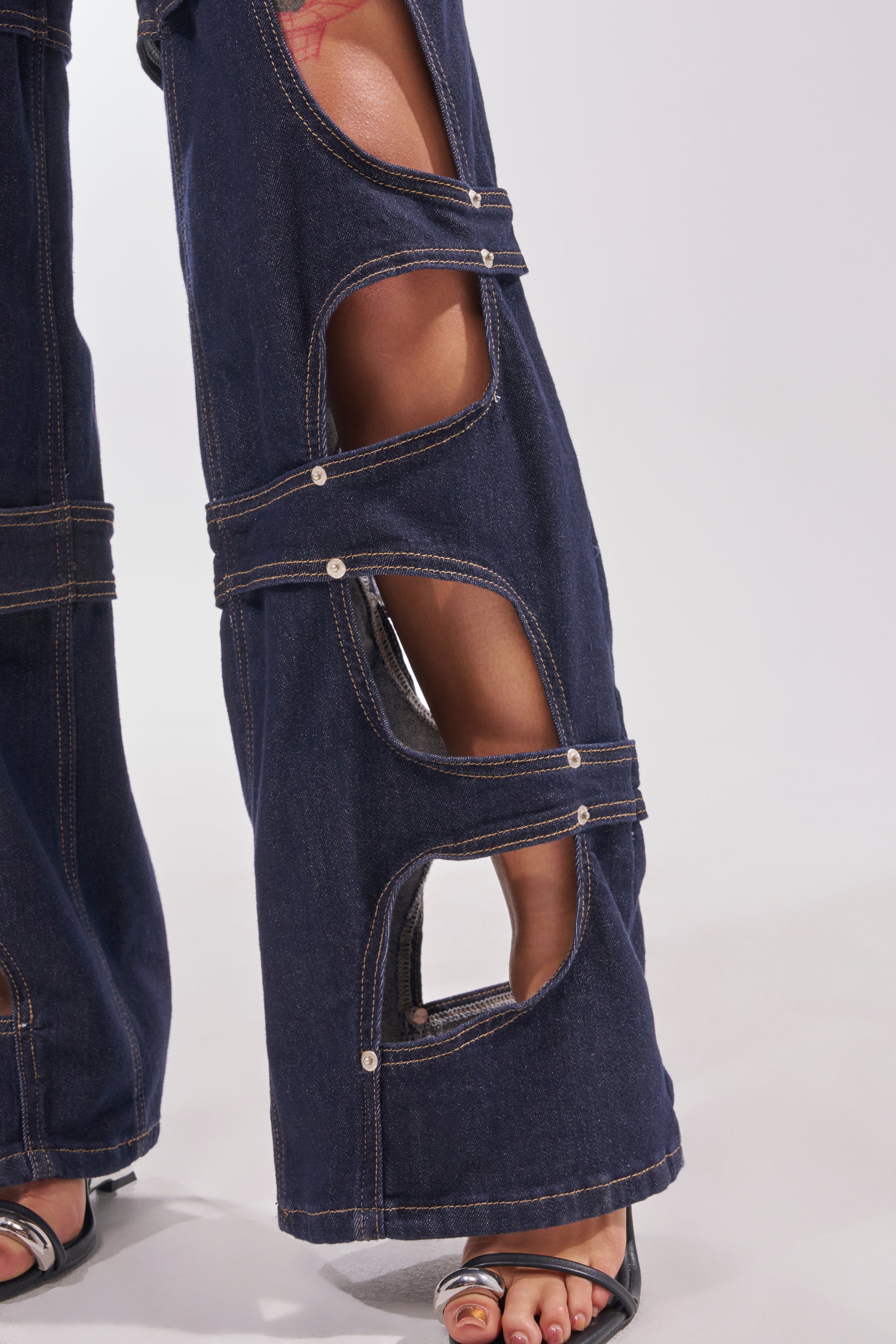 Close-up of a person wearing the BRING THE ENERGY CUT OUT DENIM JUMPSUIT, featuring a dark-wash design with bold cutouts connected by horizontal straps and buttons, flared legs, and styled with open-toed heeled sandals. 5