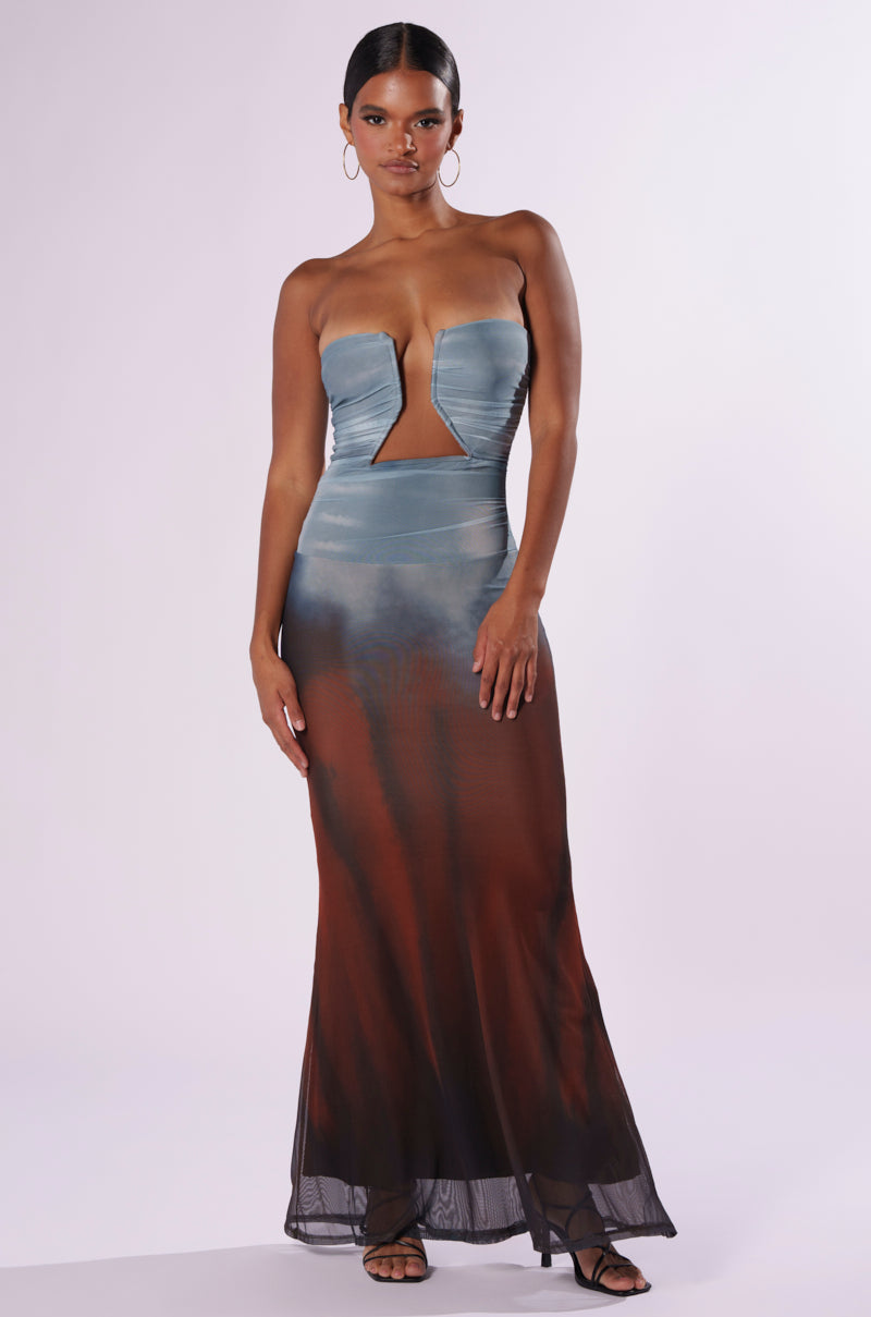 A woman is adorned in the MORNING DEW MAXI DRESS, a strapless masterpiece distinguished by an abstract ombre pattern. The gradient elegantly transitions from light blue at the top to dark brown at the bottom, and it features a distinctive chest cutout. She completes her chic ensemble with hoop earrings and black strappy heels.