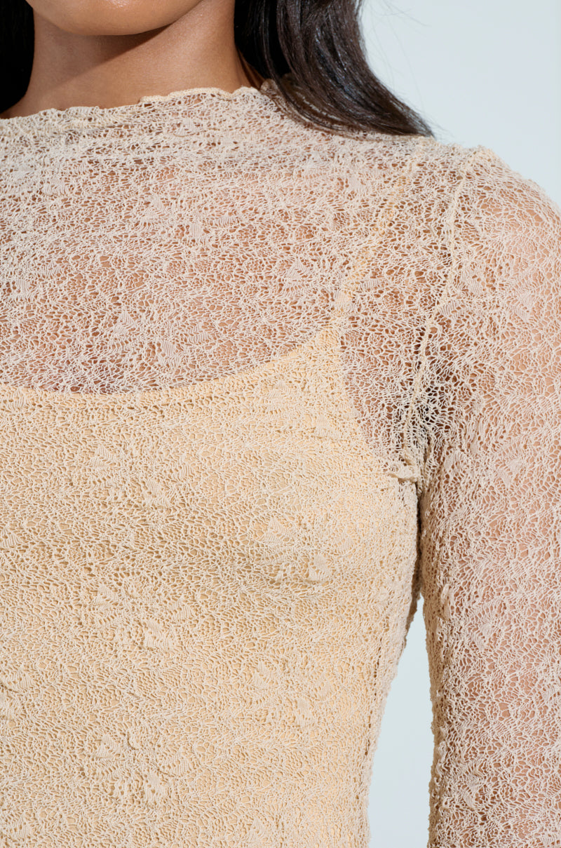 Close-up of a person wearing the "LOST IN THE CITY MAXI DRESS," a beige lace piece reminiscent of SATC style, with intricate patterns, high neckline, and long sleeves. The person's face is hidden, enhancing the mysterious vibe.