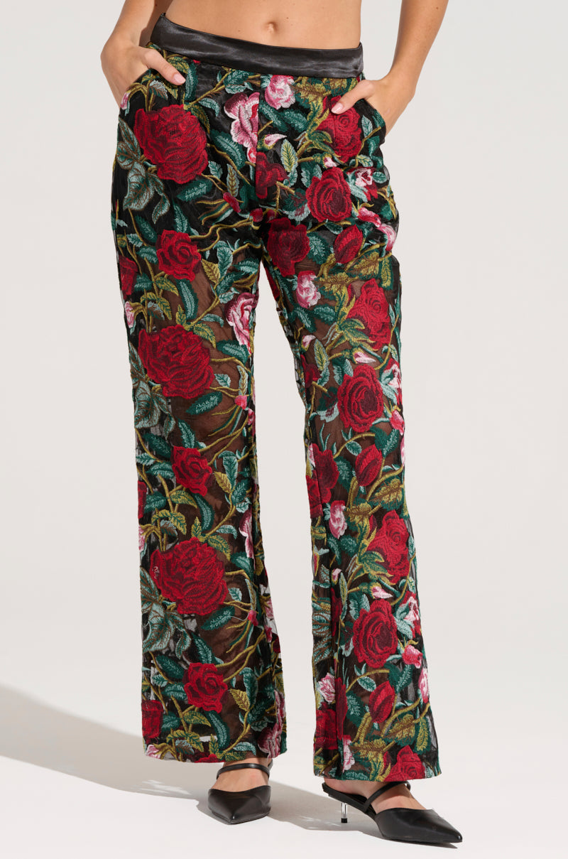 A person is modeling the EDEN LACE FLARE PANT from AKIRA Label, showcasing a vibrant embroidered floral pattern with red roses and green leaves. The pants feature a black waistband, and the individual has their hands in the pockets while wearing black pointed-toe shoes.