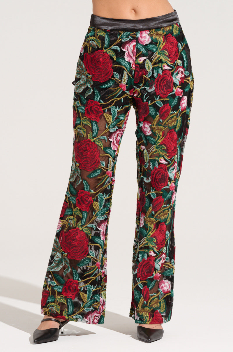 A person is wearing high-waisted EDEN LACE FLARE PANTS from the AKIRA Label, featuring a vibrant embroidered floral pattern with large red roses and greenery. The pants are paired with black pointed-toe shoes. The upper body is cropped out of the image.