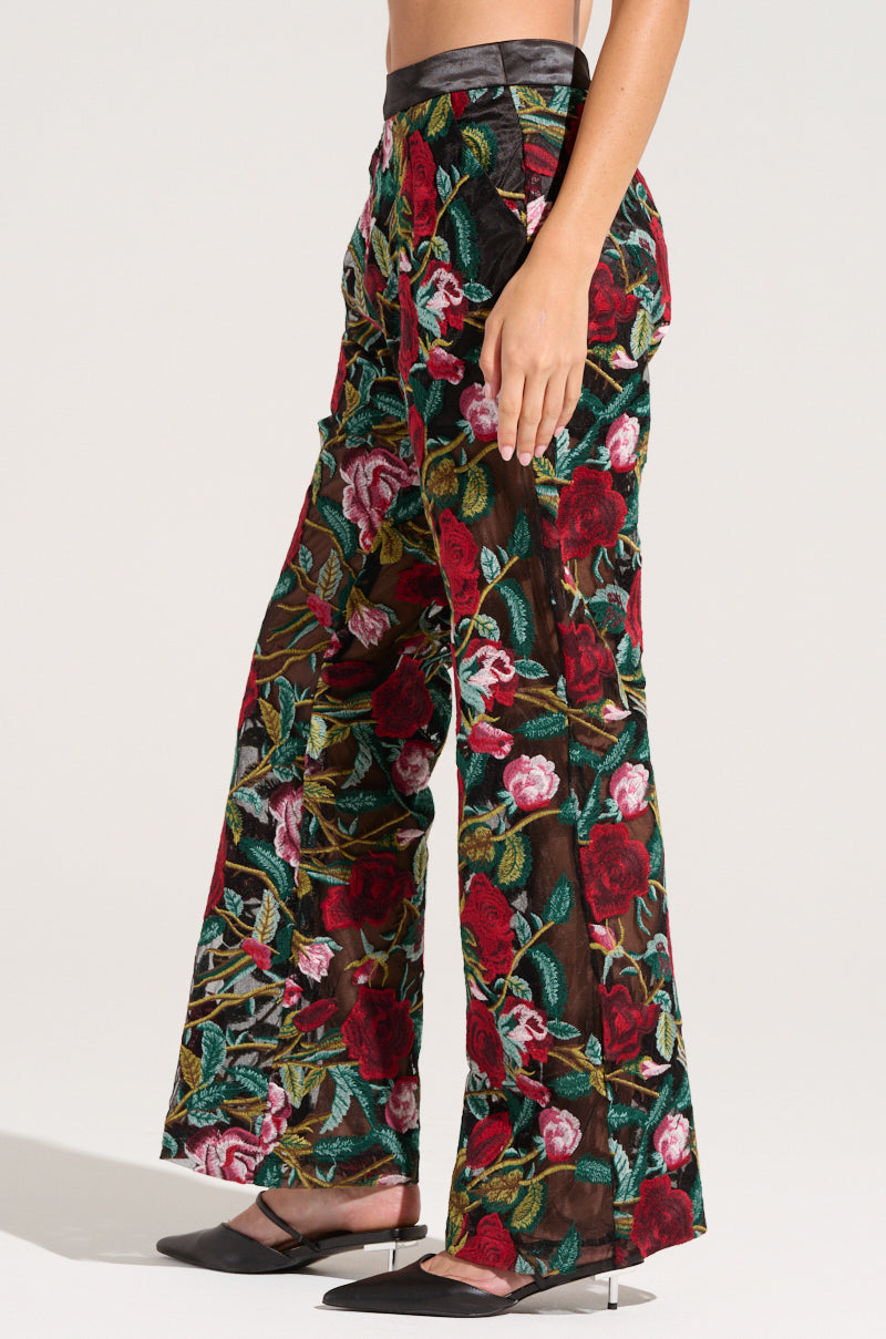 A person with bare arms is wearing high-waisted, wide-leg Eden Lace Flare Pant embellished with a vibrant embroidered floral pattern in shades of red, pink, and green. They are also sporting black pointed-toe shoes. The background is plain and well-lit.