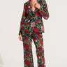 A woman stands confidently against a plain white background, sporting the AKIRA Label's EDEN LACE FLARE PANT, featuring a vibrant floral design with red roses and green leaves. She accessorizes with large dark sunglasses and black pointed-toe shoes. Her hair is pulled back, and her right hand touches her collar, showcasing the intricate embroidered pattern.