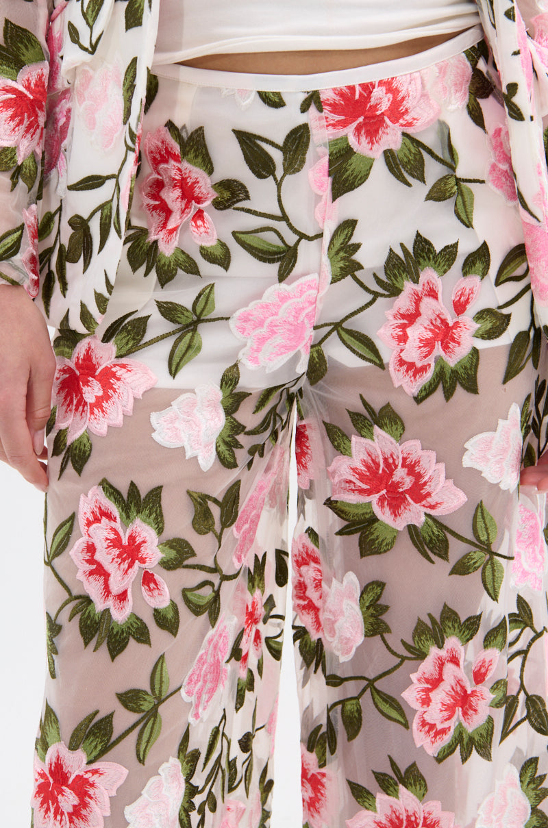 A close-up of a person wearing BLOSSOM LACE PALAZZO PANTS, showcasing a white base with bold pink and red floral lace embroidery and green leaves. The matching jacket enhances the coordinated, vibrant wide-leg silhouette.