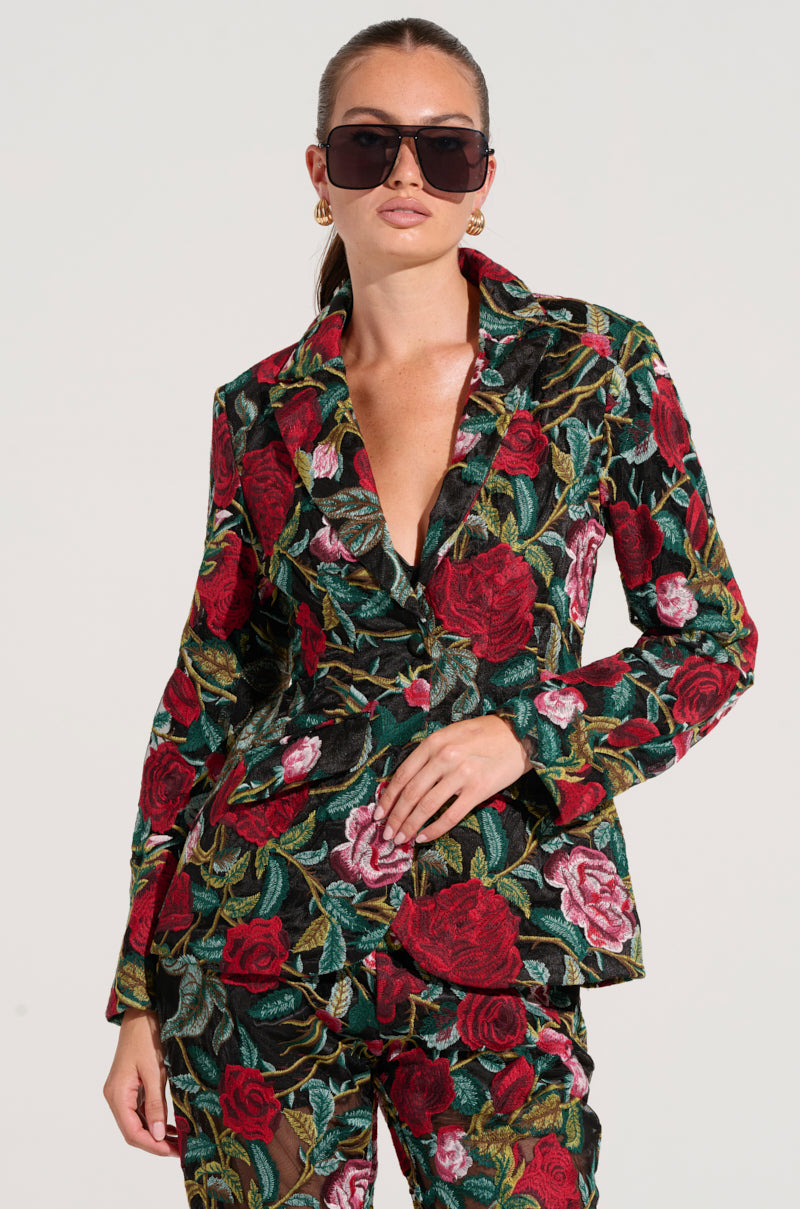 A person wearing the EDEN LACE BLAZER, adorned with an embroidered floral pattern of red and pink roses against a dark green backdrop, is posing confidently with one hand on their waist. They are also sporting dark sunglasses and gold earrings, standing against a plain white background.