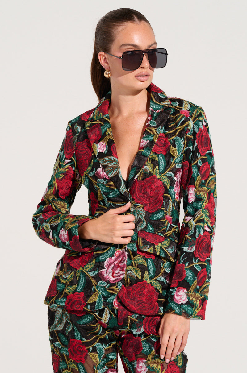 A woman wearing oversized black sunglasses and the EDEN LACE BLAZER, which is elegantly adorned with a fashionable embroidered floral pattern in vibrant red, pink, and green hues. Her hair is styled into a sleek ponytail, complemented by gold earrings. She exudes confidence in her fitted longline silhouette against a plain background.