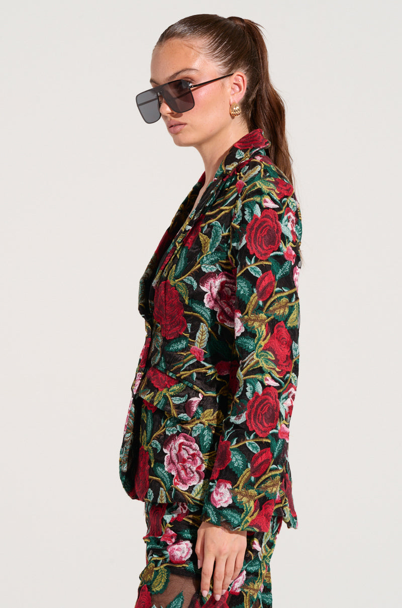 A person with long, pulled-back hair is wearing oversized sunglasses, gold earrings, and an EDEN LACE BLAZER that forms part of a matching suit adorned with vibrant red and pink roses. The fitted longline silhouette enhances their elegant appearance as they face right against a plain background.