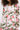 A person wears the BLOSSOM LACE BLAZER, a structured fit white floral lace blazer with sheer sleeves in vibrant spring hues, styled with a chunky gold necklace. Their face is partially out of frame.