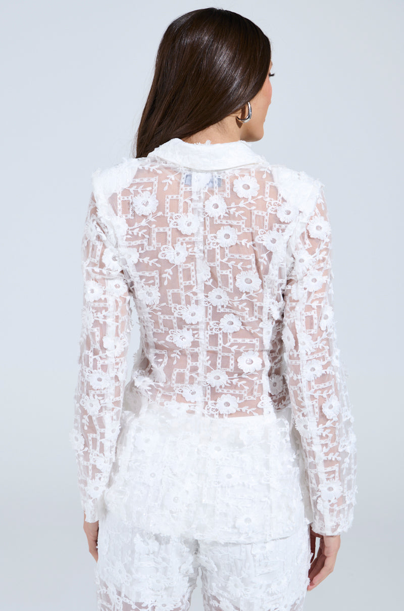 A woman with long brown hair is turned away, wearing a sheer white lace long-sleeve top and matching floral-patterned pants, reminiscent of an elegant CELEBRATION TIME BLAZER. The background is plain and light gray.