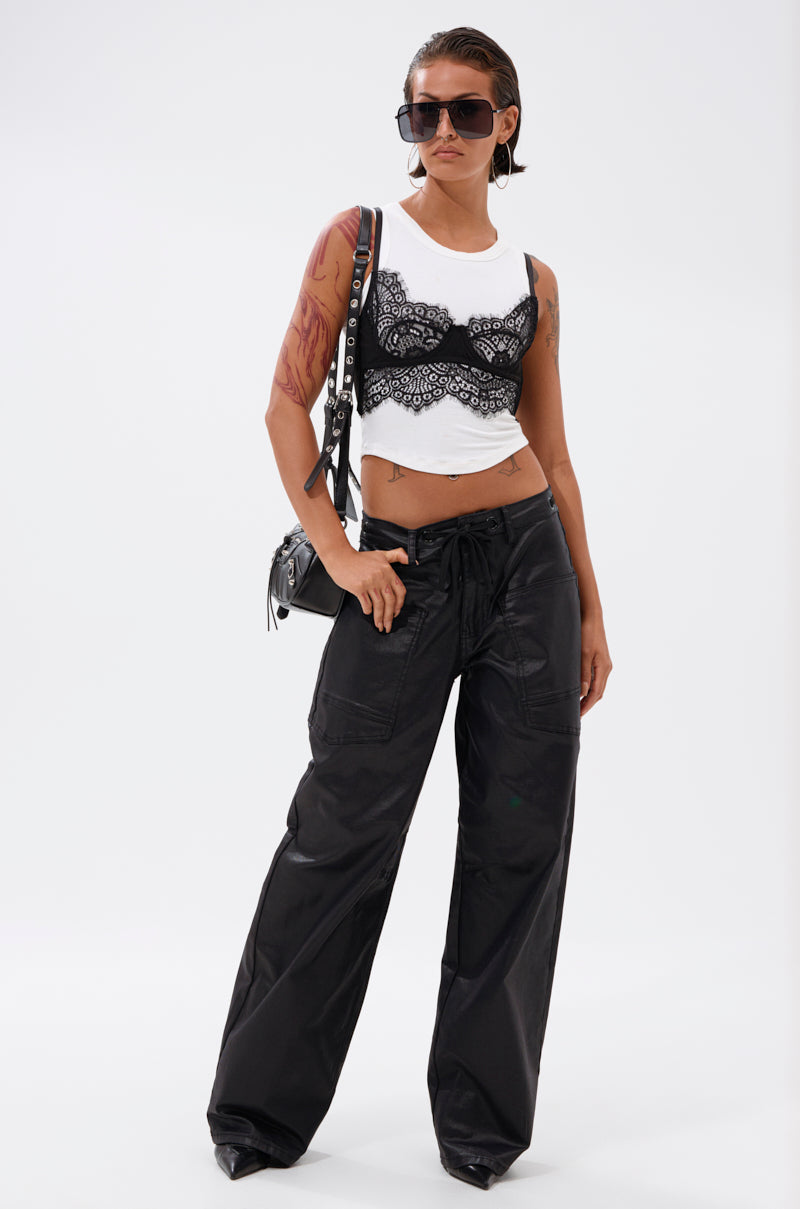 A person with short hair and sunglasses poses against a plain white background wearing a white crop top, black lace bralette, DONT GIVE UP COATED WIDE LEG DENIM WITH GROMMET, and a black shoulder bag.