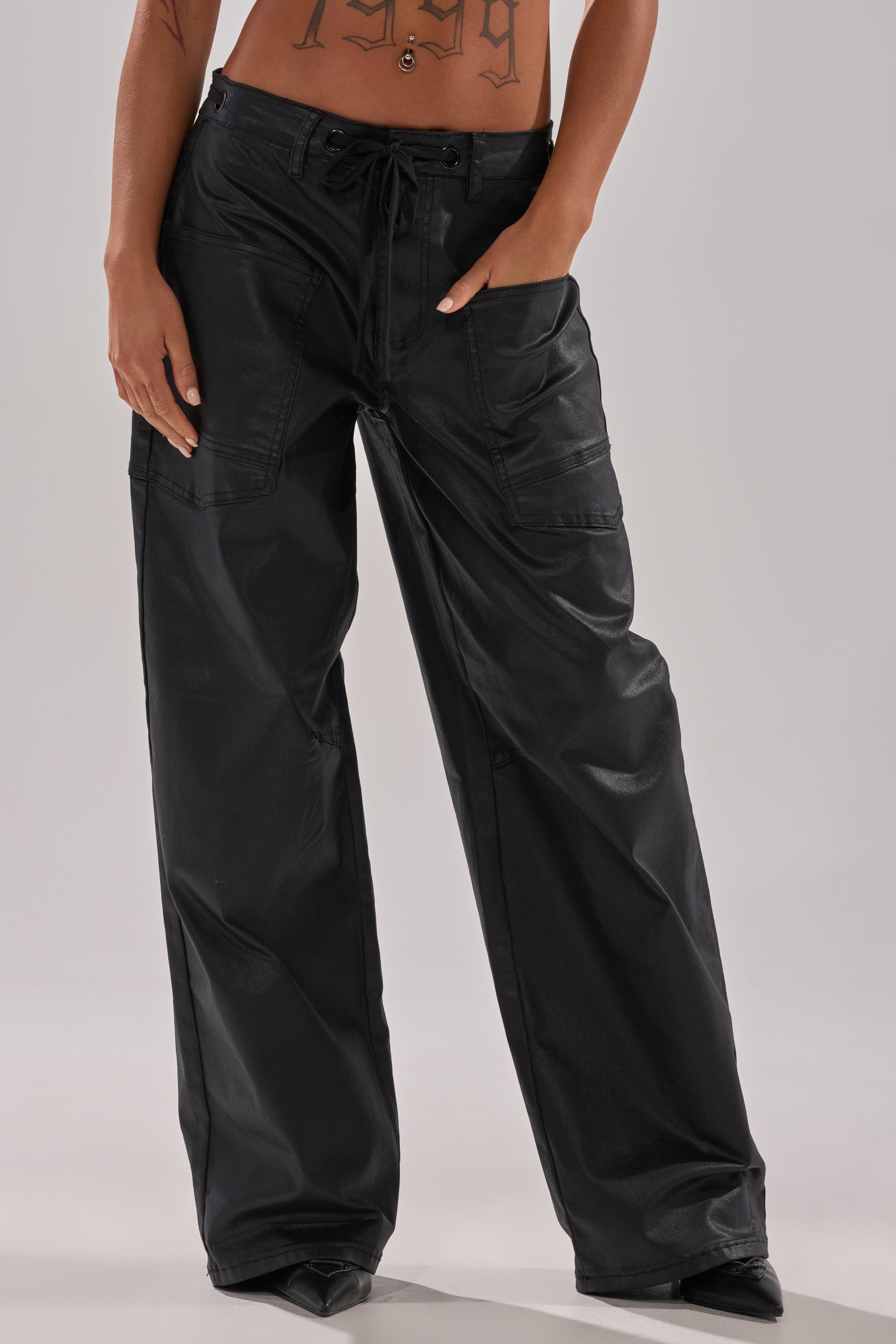 Wearing the DONT GIVE UP COATED WIDE LEG DENIM WITH GROMMET—black, loose street-style jeans with a shiny coated finish, grommet details, multiple pockets, and a drawstring waist. Midriff and tattoos are visible.