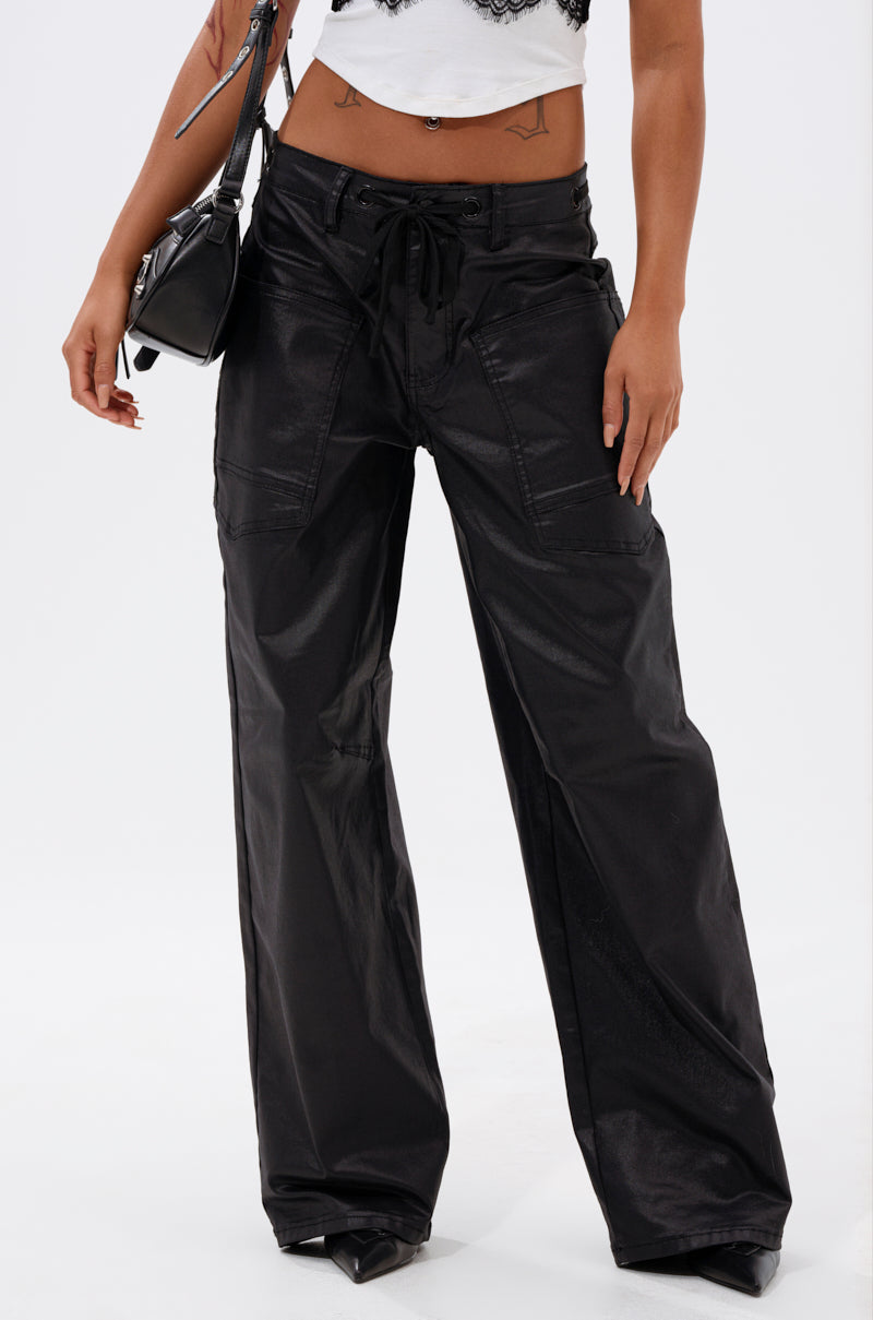 A person wearing DONT GIVE UP COATED WIDE LEG DENIM WITH GROMMET—shiny black wide-leg denim cargo pants with front pockets and a drawstring waist—is shown from the lower torso and legs.