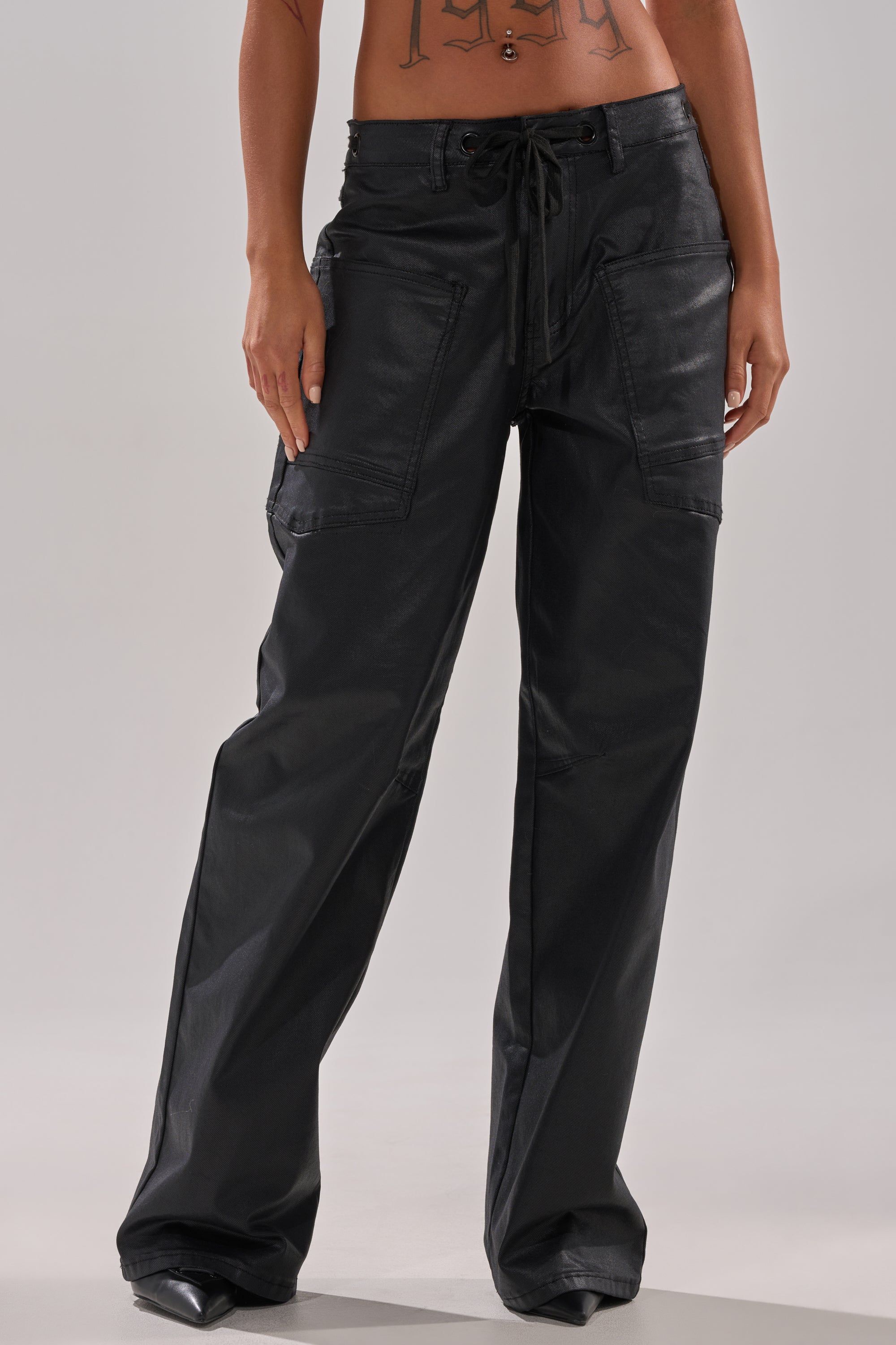 A person wears DONT GIVE UP COATED WIDE LEG DENIM WITH GROMMET, featuring a loose street-style fit and large front pockets. Only the lower torso and legs are shown against a plain background, emphasizing these urban-inspired pants' relaxed vibe.