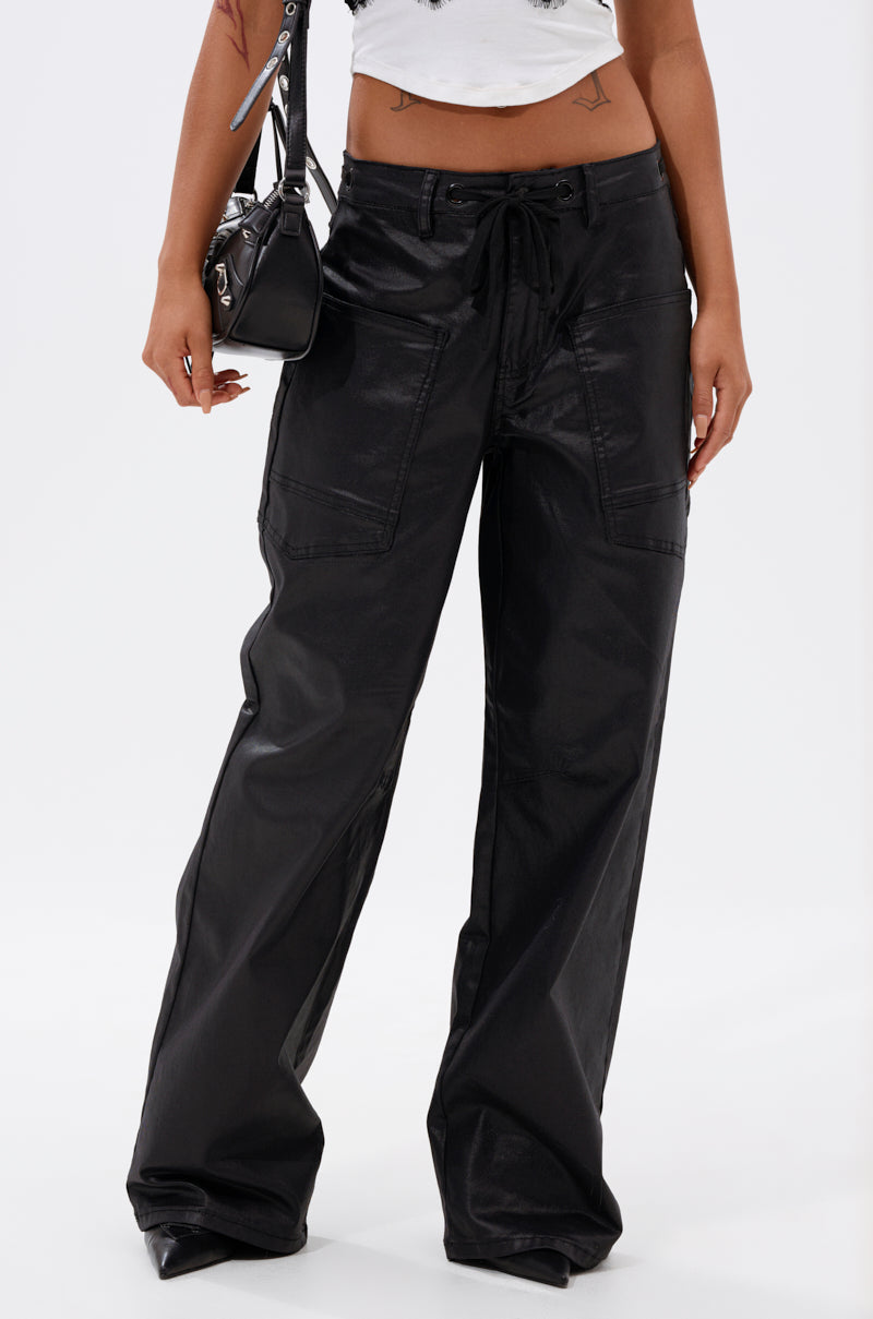 A person wearing the DONT GIVE UP COATED WIDE LEG DENIM WITH GROMMET, featuring a waxed finish and large front pockets, paired with a white crop top and carrying a small black shoulder bag. Only the lower half of their body is visible.