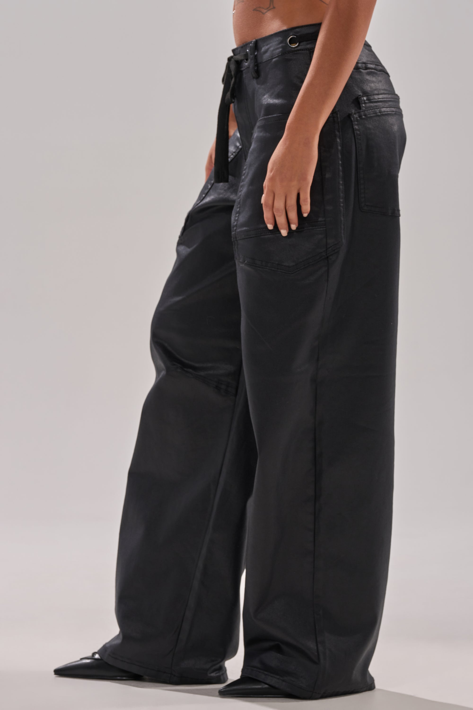 Wearing the DONT GIVE UP COATED WIDE LEG DENIM WITH GROMMET, featuring a shiny coated finish, high-waisted fit, large front pockets, and slight flare at the hem, shown waist down against a plain background.