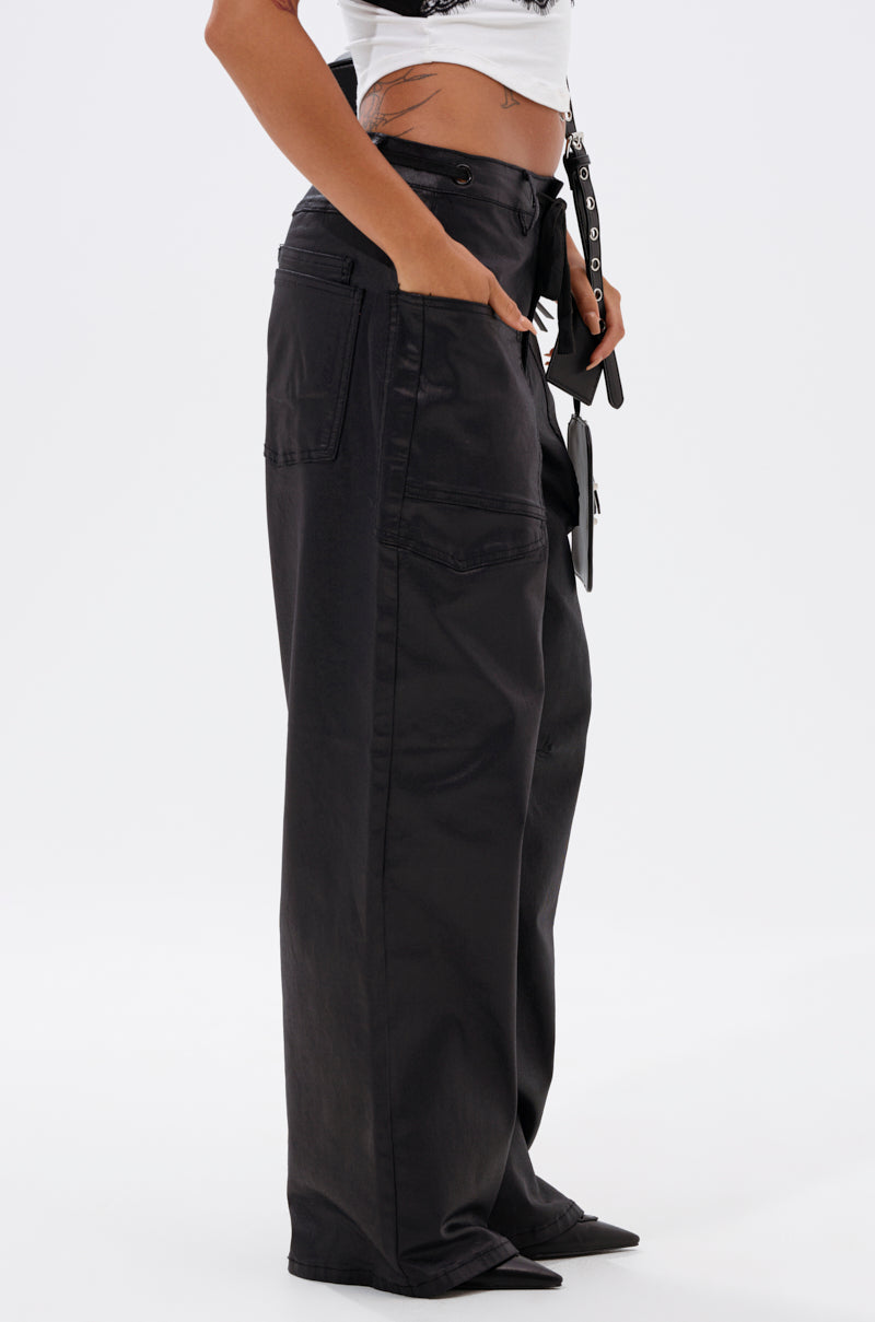 Someone wearing the DONT GIVE UP COATED WIDE LEG DENIM WITH GROMMET, hands in pockets, paired with a white crop top. Side view against a plain white background.