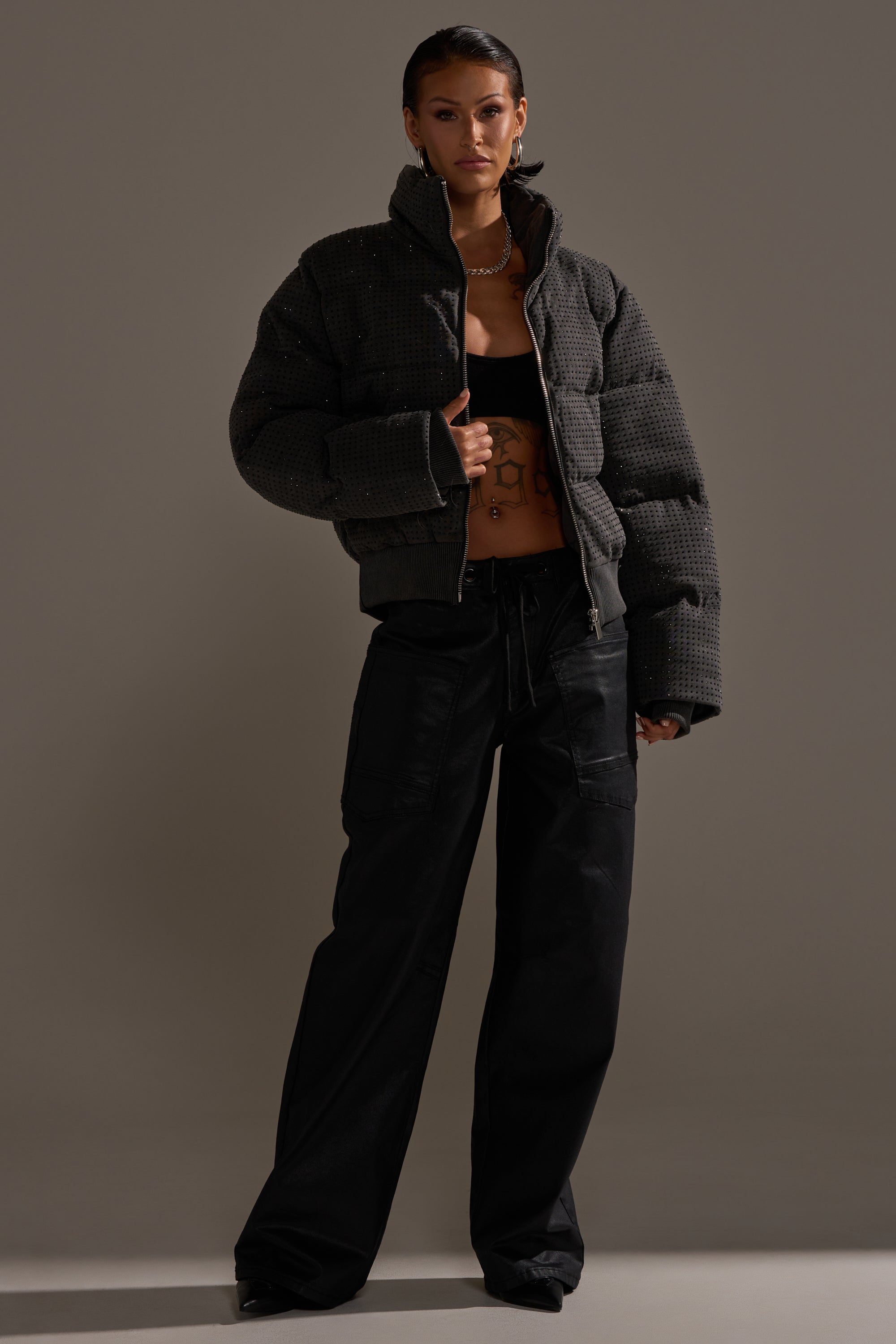 A woman poses confidently against a plain backdrop wearing the DONT GIVE UP COATED WIDE LEG DENIM WITH GROMMET, a textured dark puffer jacket, and a black crop top. She has short hair and hoop earrings, showcasing effortless style.