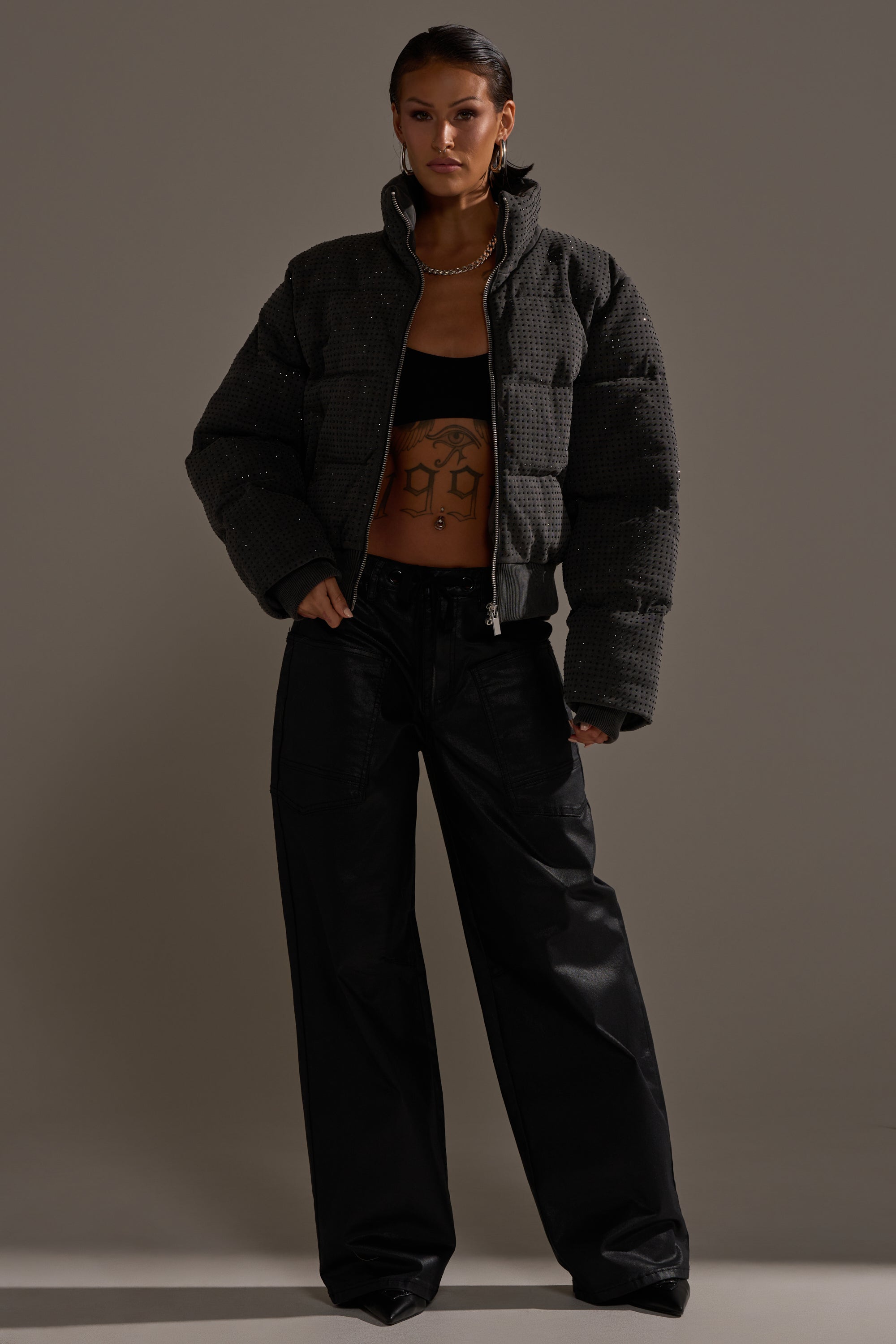 A woman stands confidently in a dark gray puffer jacket, black crop top, layered necklaces, and DONT GIVE UP COATED WIDE LEG DENIM WITH GROMMET, showcasing her chest tattoo against a neutral background.