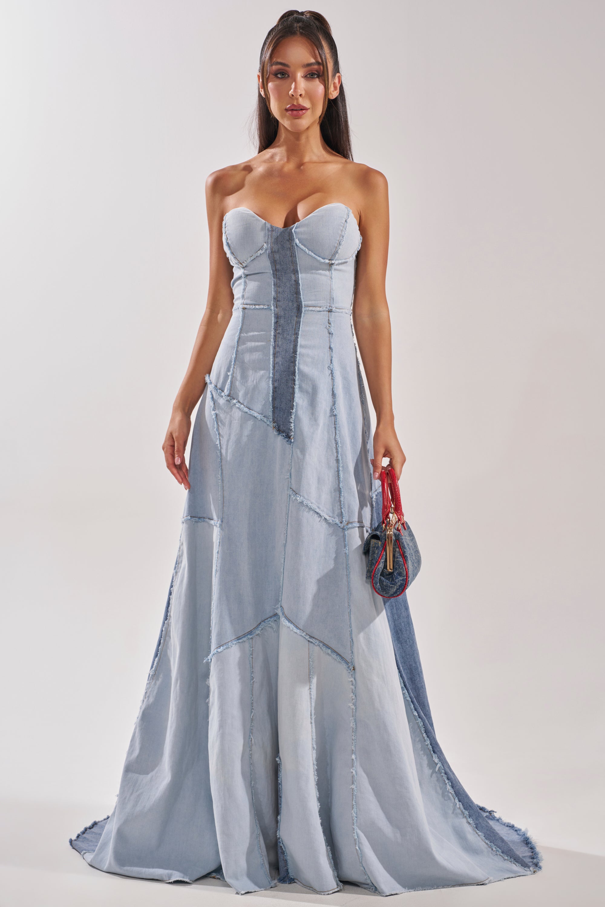 A woman wears the IT'S BRITNEY PATCHWORK DENIM MAXI DRESS, featuring strapless light and dark blue denim panels. She holds a small handbag, her long dark hair down, against a plain light-colored background. 0