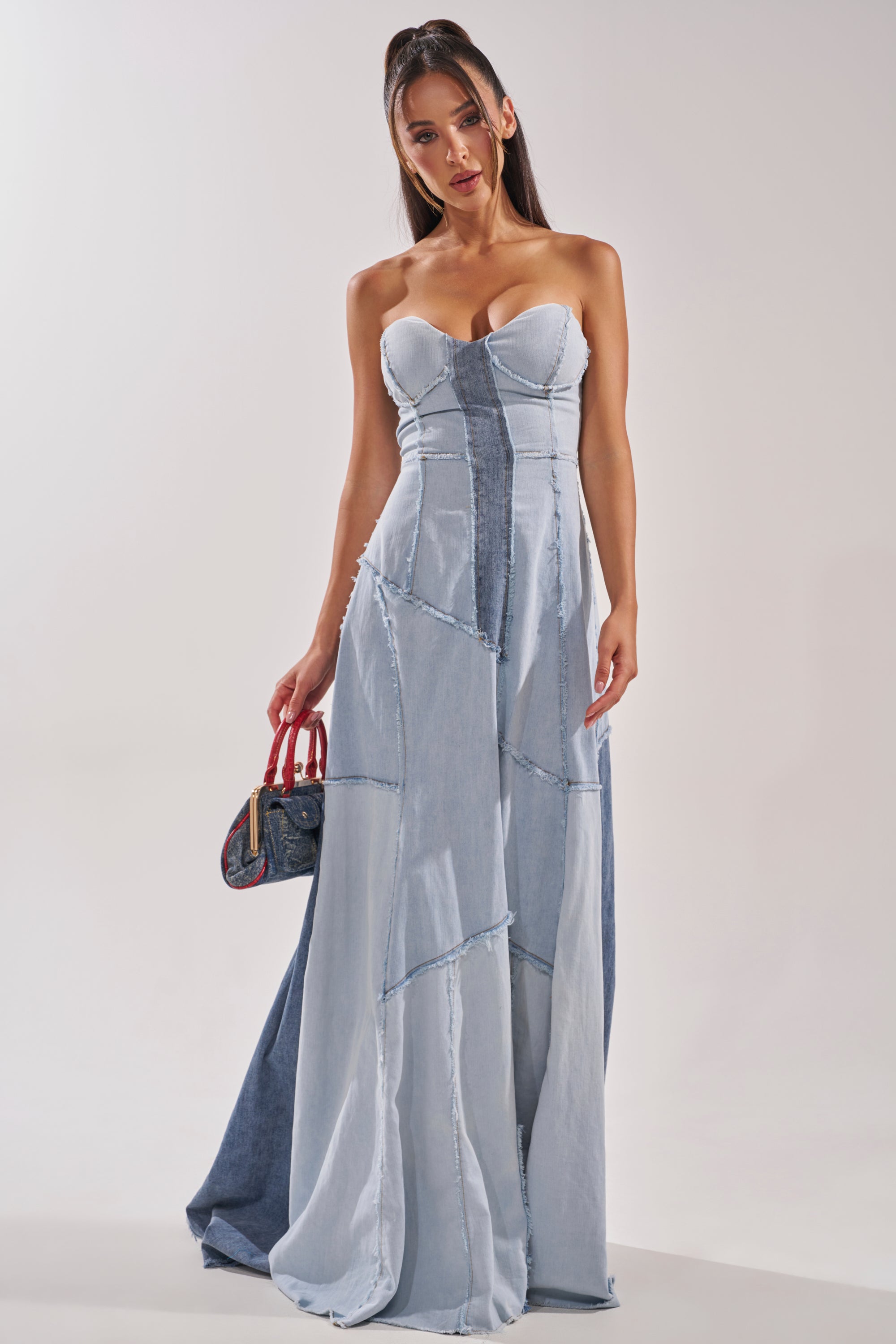 A woman in the IT'S BRITNEY PATCHWORK DENIM MAXI DRESS stands against a plain background, holding a small handbag with red handles. Her long hair is half-up, and she gazes confidently at the camera. 1