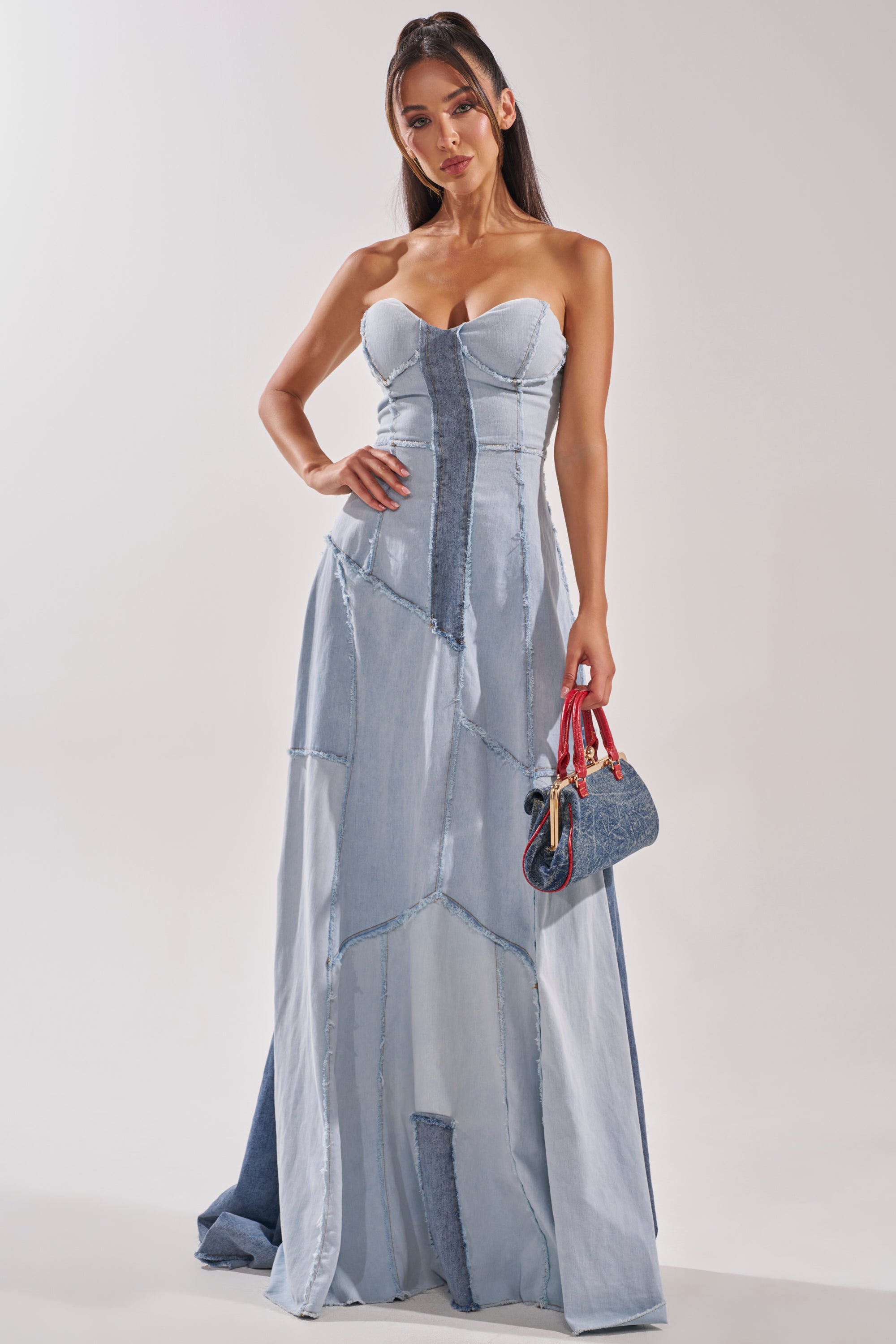 A woman poses against a plain background in the IT'S BRITNEY PATCHWORK DENIM MAXI DRESS, a strapless, floor-length maxi. She holds a small handbag with colorful straps and wears her long, straight hair down. 2